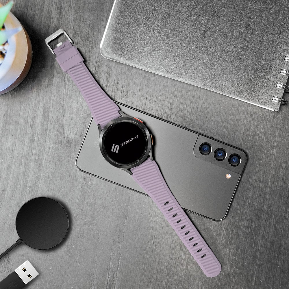 Eine lavendelfarbene Smartwatch mit passendem Armband liegt auf einem grauen Telefon. Ein Ladepad und ein Laptop sind ebenfalls vorhanden.