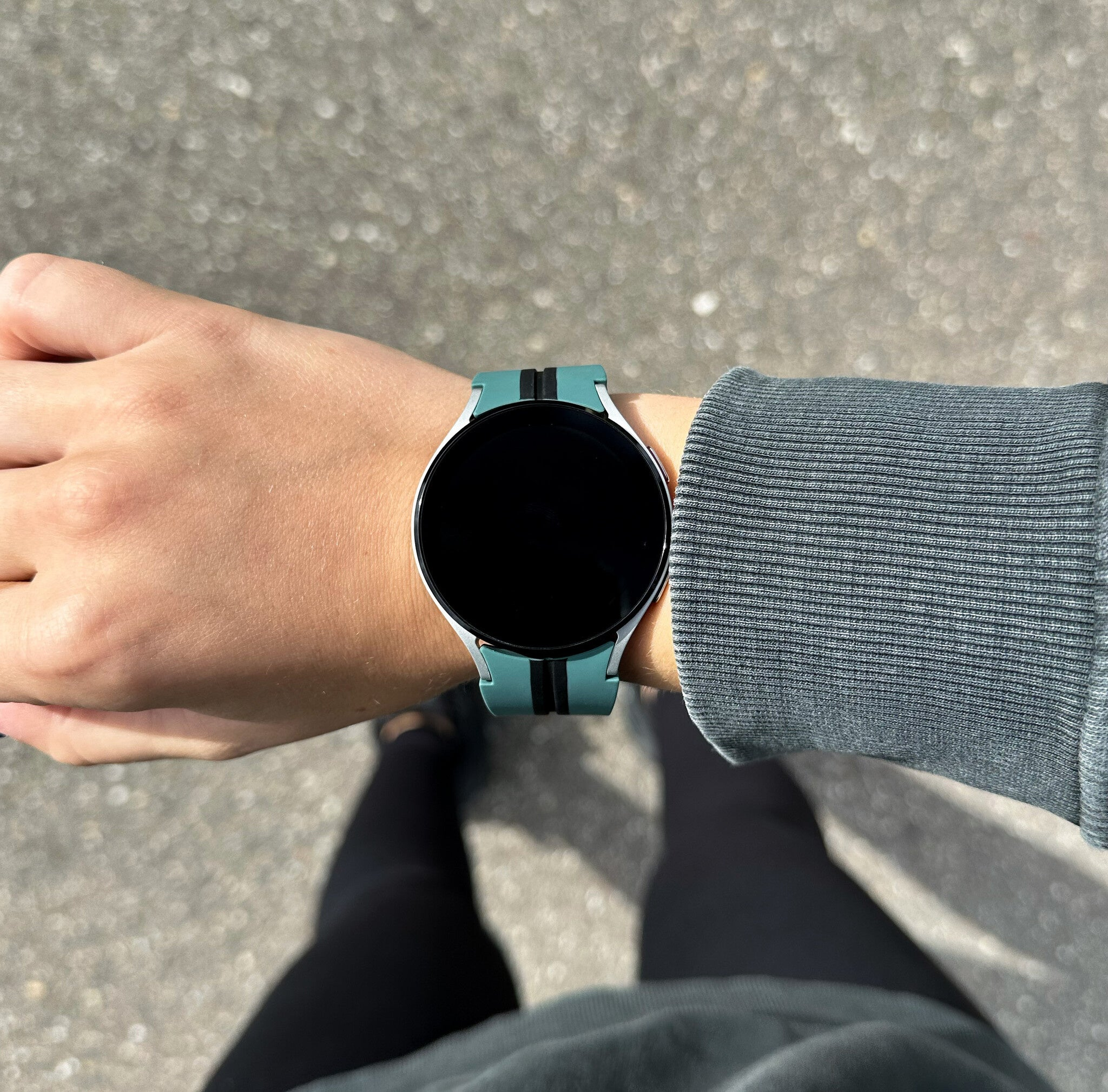 Das Handgelenk einer Person mit einer teal Smartwatch. Die Uhr hat einen schwarzen Bildschirm und eine graue Jacke ist sichtbar.