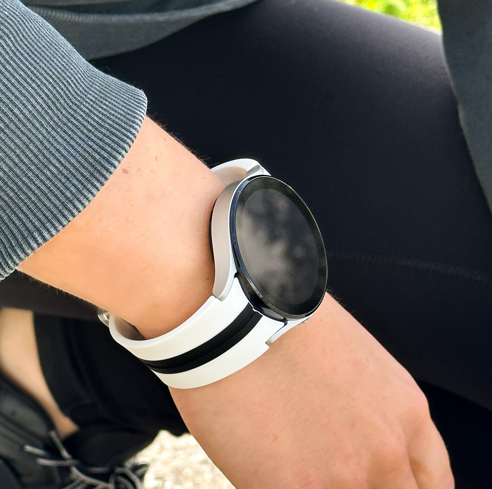 Das Handgelenk einer Person mit einer weißen Smartwatch. Die Uhr hat einen schwarzen Bildschirm und schwarz-weiße Armbänder. Person trägt grauen Ärmel.