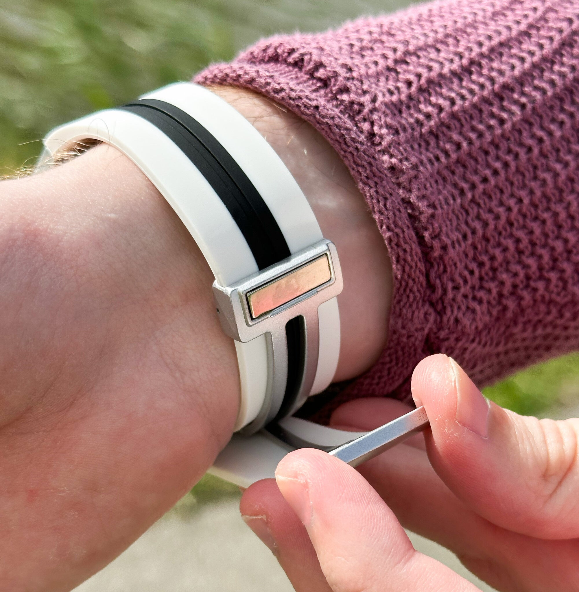 Das Handgelenk einer Person mit einer weiß-schwarz gestreiften Uhr. Die Person trägt einen rosa Pullover. Eine andere Hand hält ein Metallwerkzeug.