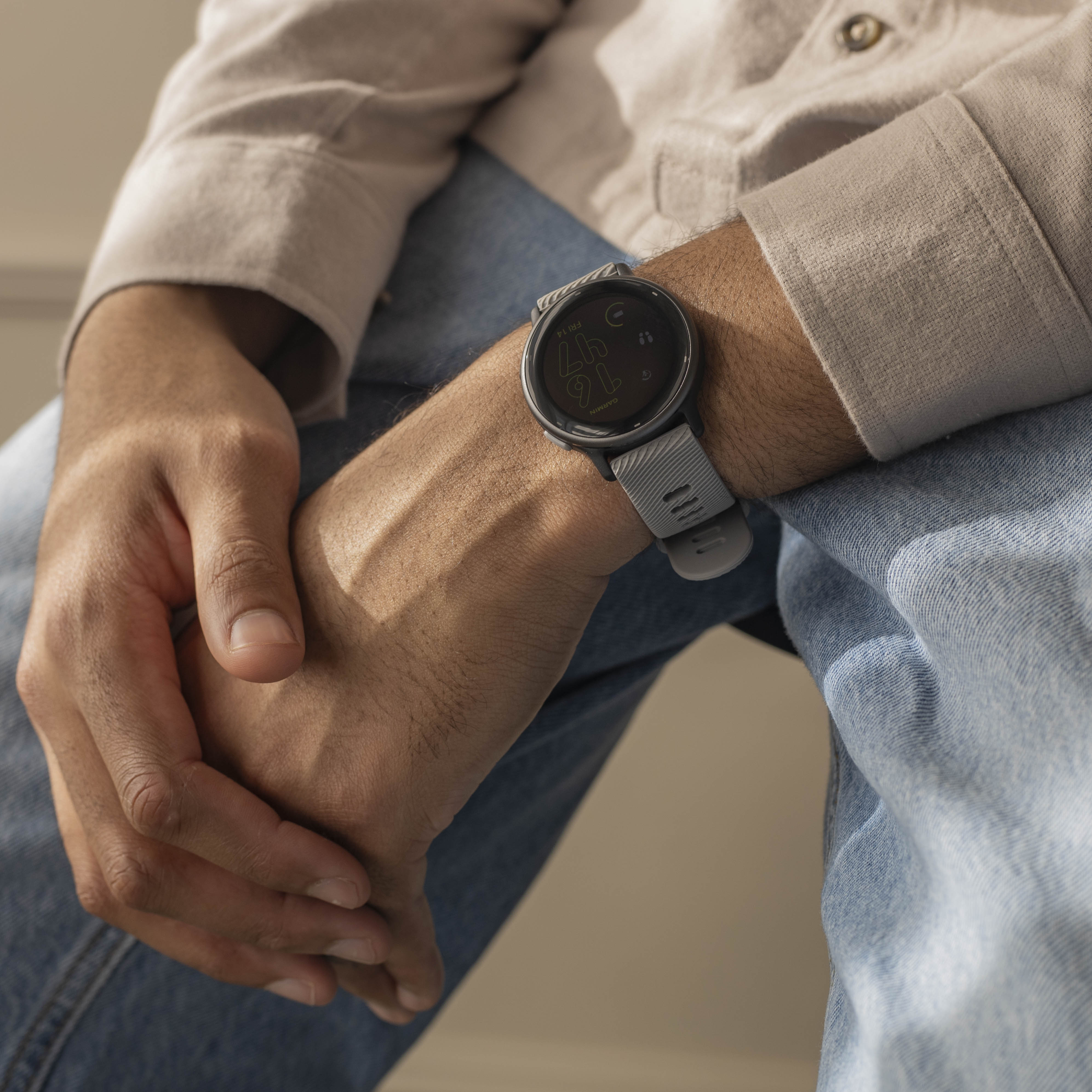 Eine Person trägt eine graue Uhr am Handgelenk. Sie trägt ein beiges Hemd und blaue Jeans, die Hände sind gefaltet.