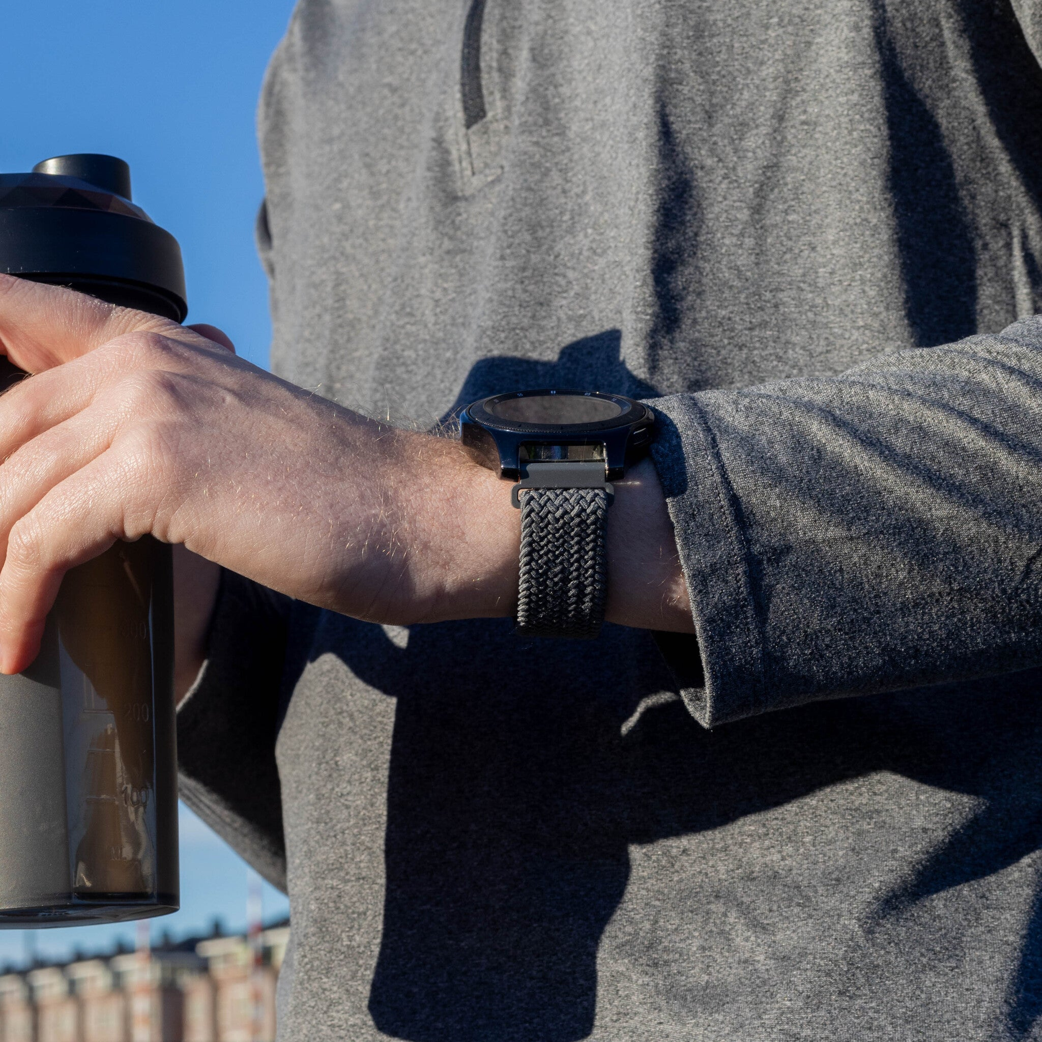 Der Arm eines Mannes hält eine Wasserflasche und trägt eine Smartwatch mit geflochtenem Armband. Blauer Himmel im Hintergrund.