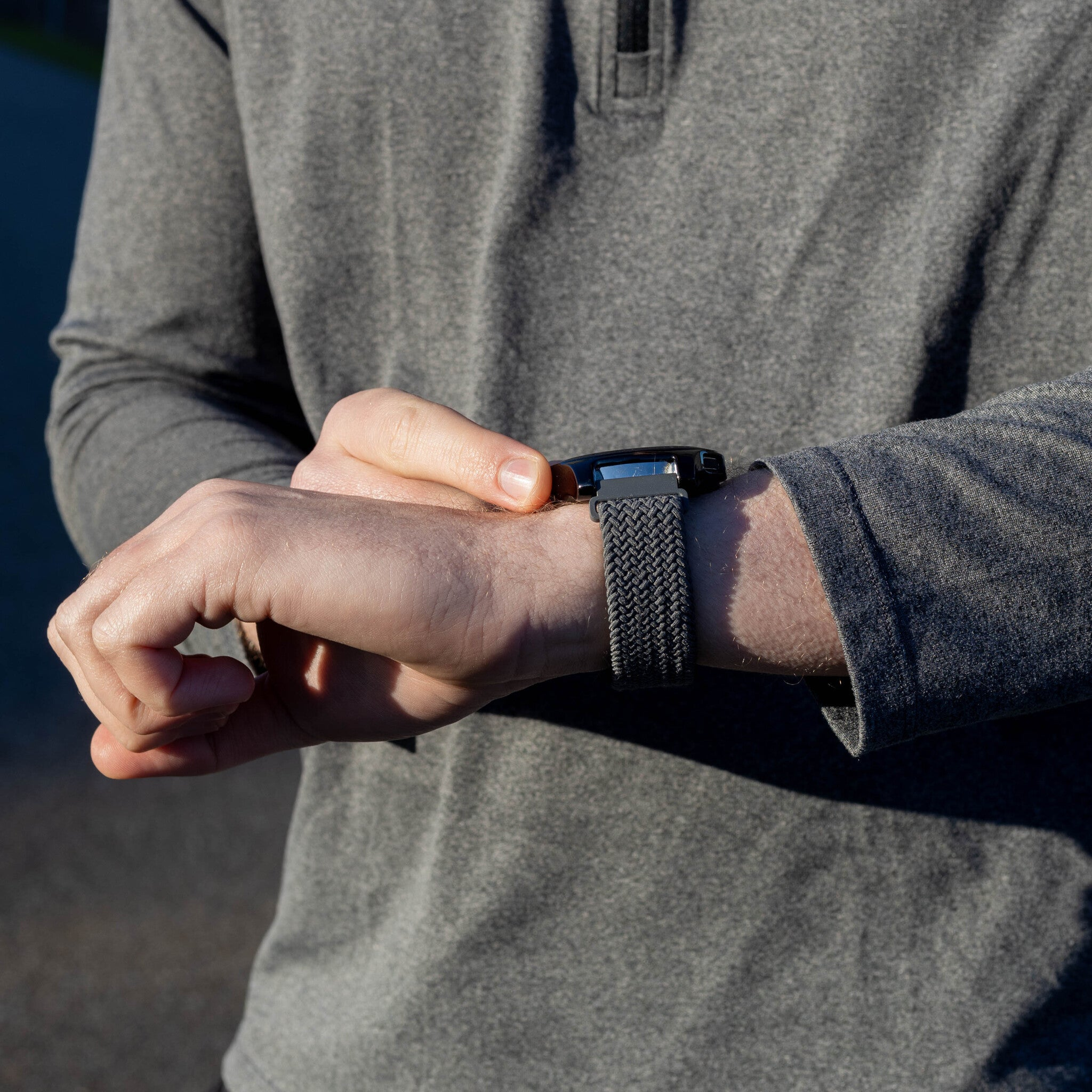 Eine Person im grauen Hemd überprüft die Uhr an ihrem Handgelenk. Die Uhr hat ein graues, gewebtes Armband und ein schwarzes Zifferblatt.