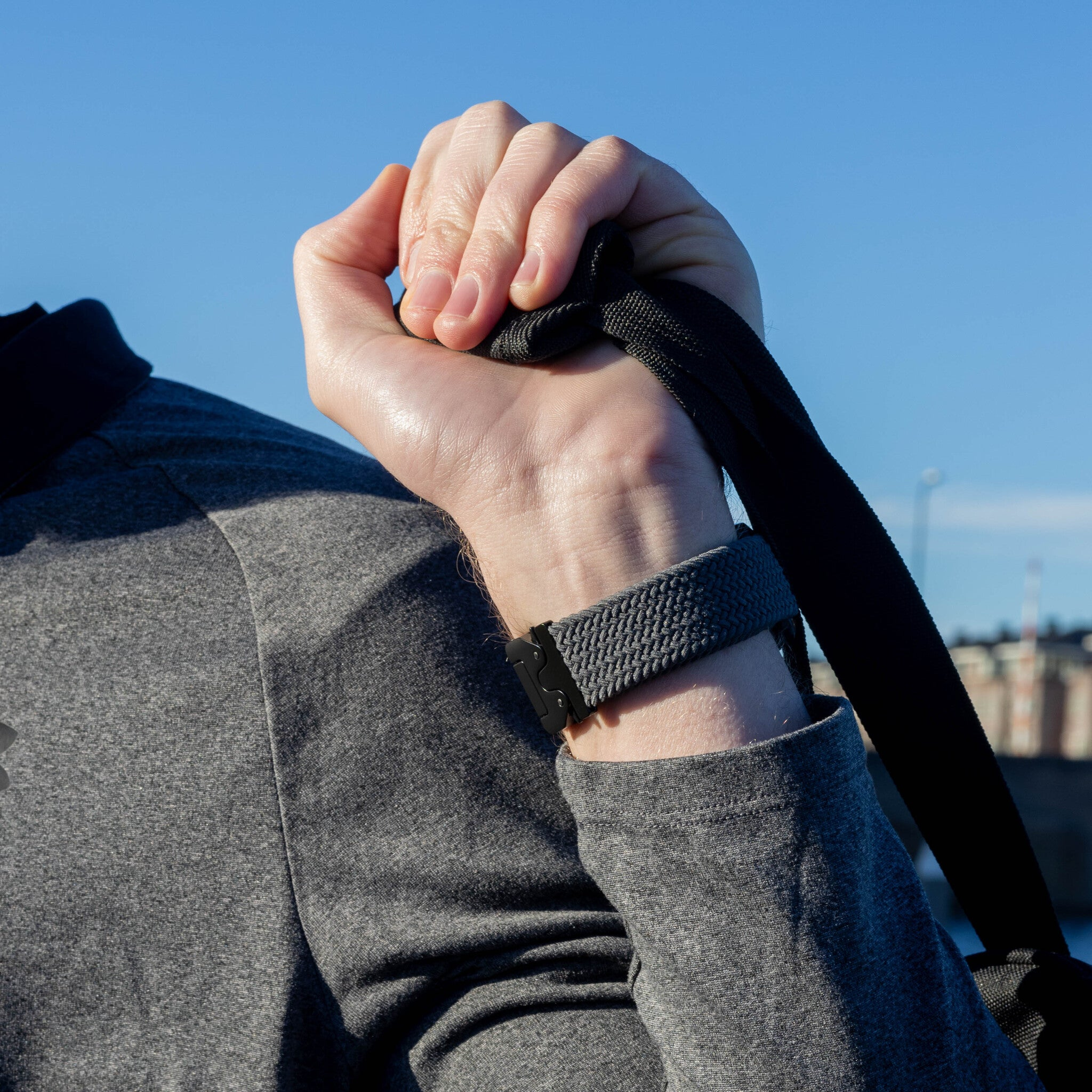 Hand hält einen schwarzen Riemen und trägt eine graue Uhr. Die Person trägt ein graues Hemd mit blauem Himmel im Hintergrund.
