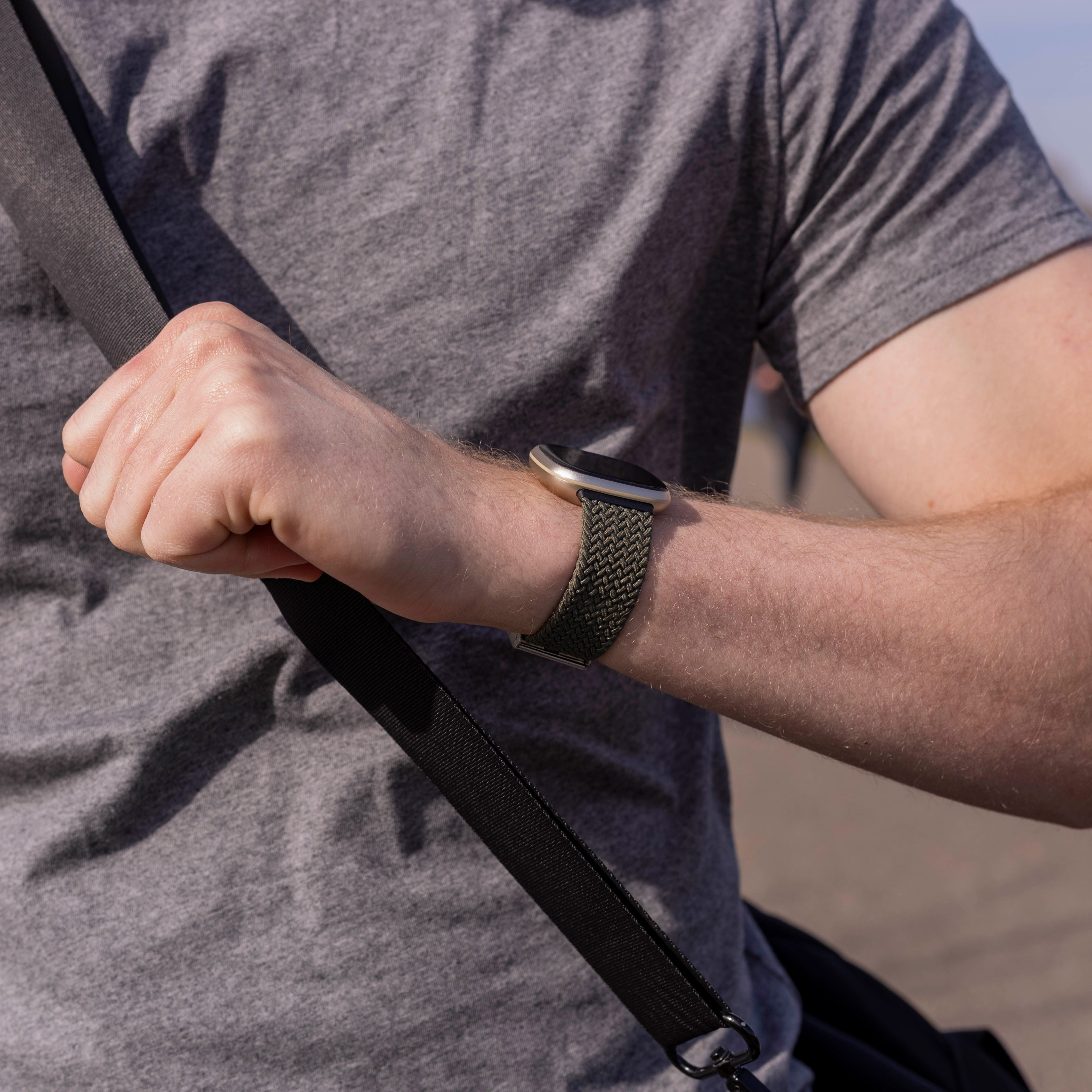 Eine Person trägt ein graues Hemd, eine Uhr mit grünem Armband und hält eine schwarze Tasche. Die Haut ist hell.