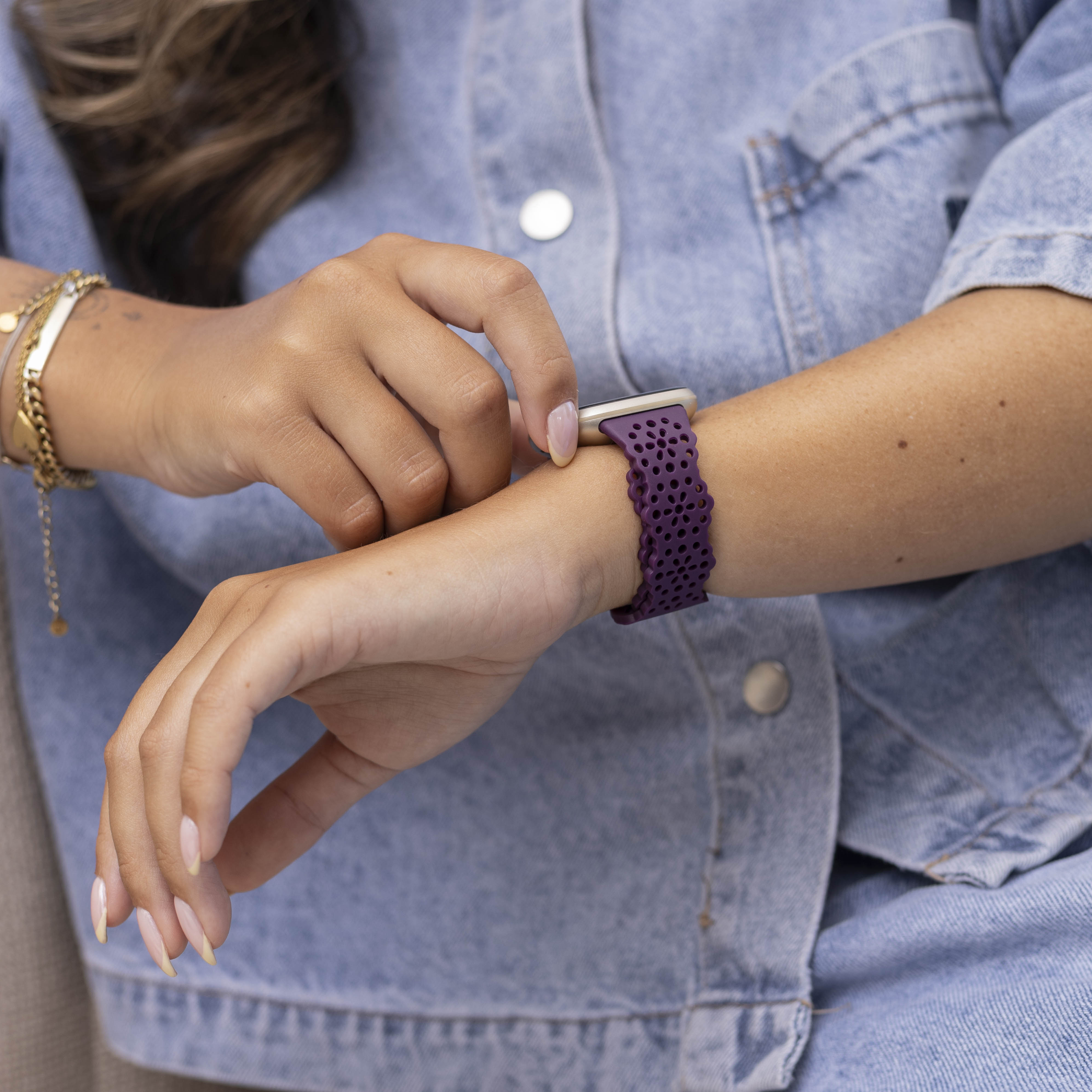 Eine Person in einer Jeansjacke stellt ein lila Uhrenarmband ein. Goldene Armbänder schmücken das linke Handgelenk. Nahaufnahme.
