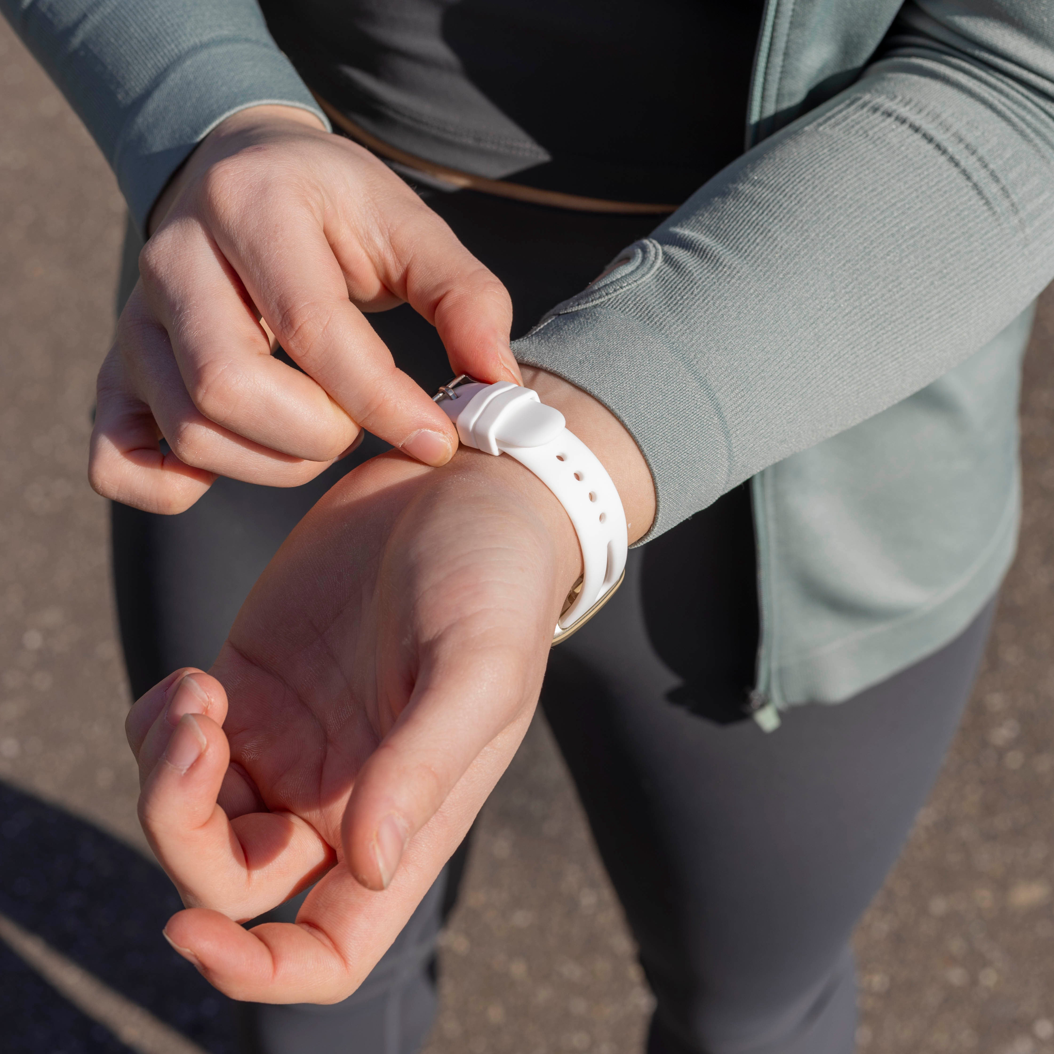 Eine Person befestigt eine weiße Uhr an ihrem Handgelenk. Sie trägt eine tealfarbene Jacke und graue Leggings.