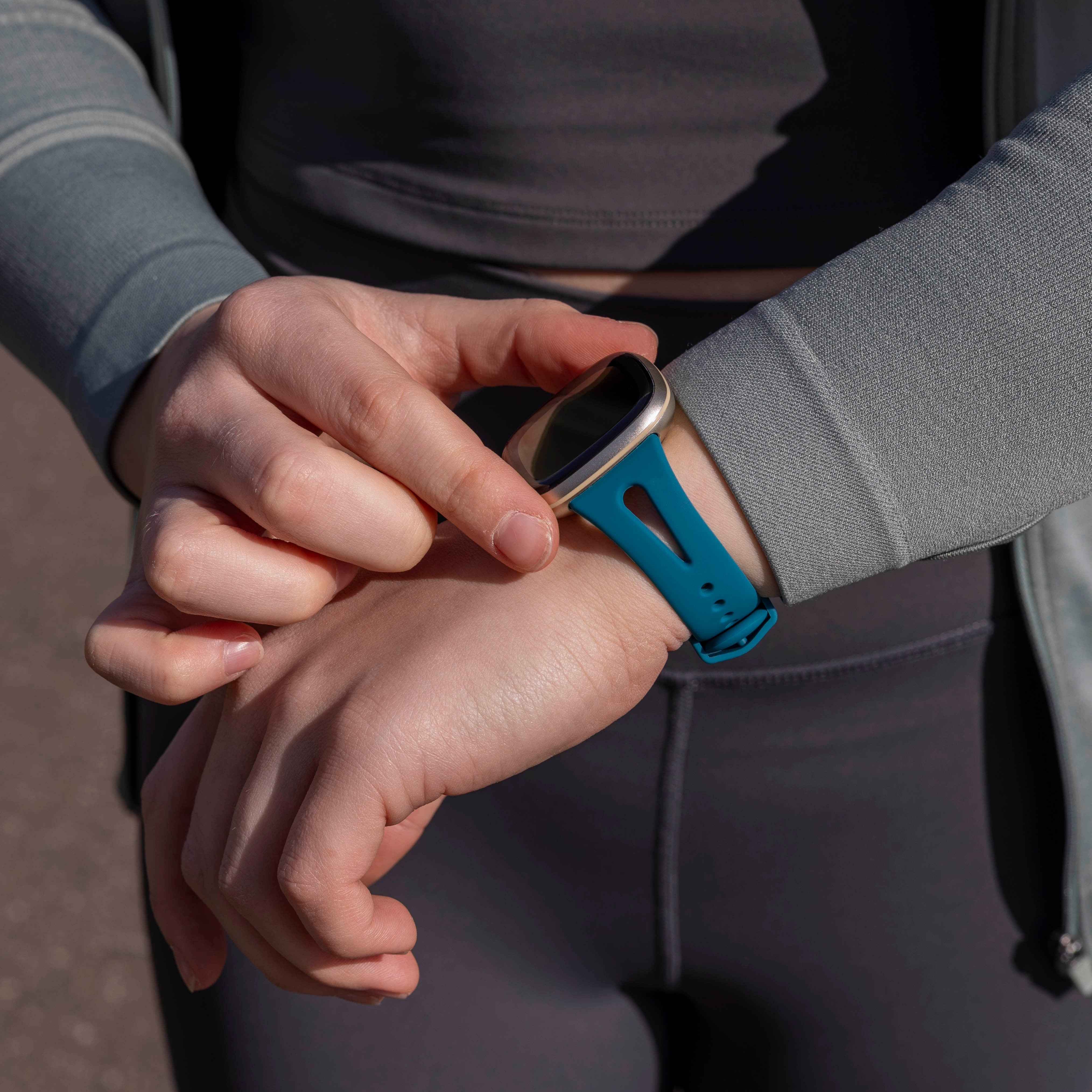 Eine Person im grauen Outfit stellt eine Smartwatch mit tealfarbenem Armband am Handgelenk ein. Der Hintergrund ist unscharf.