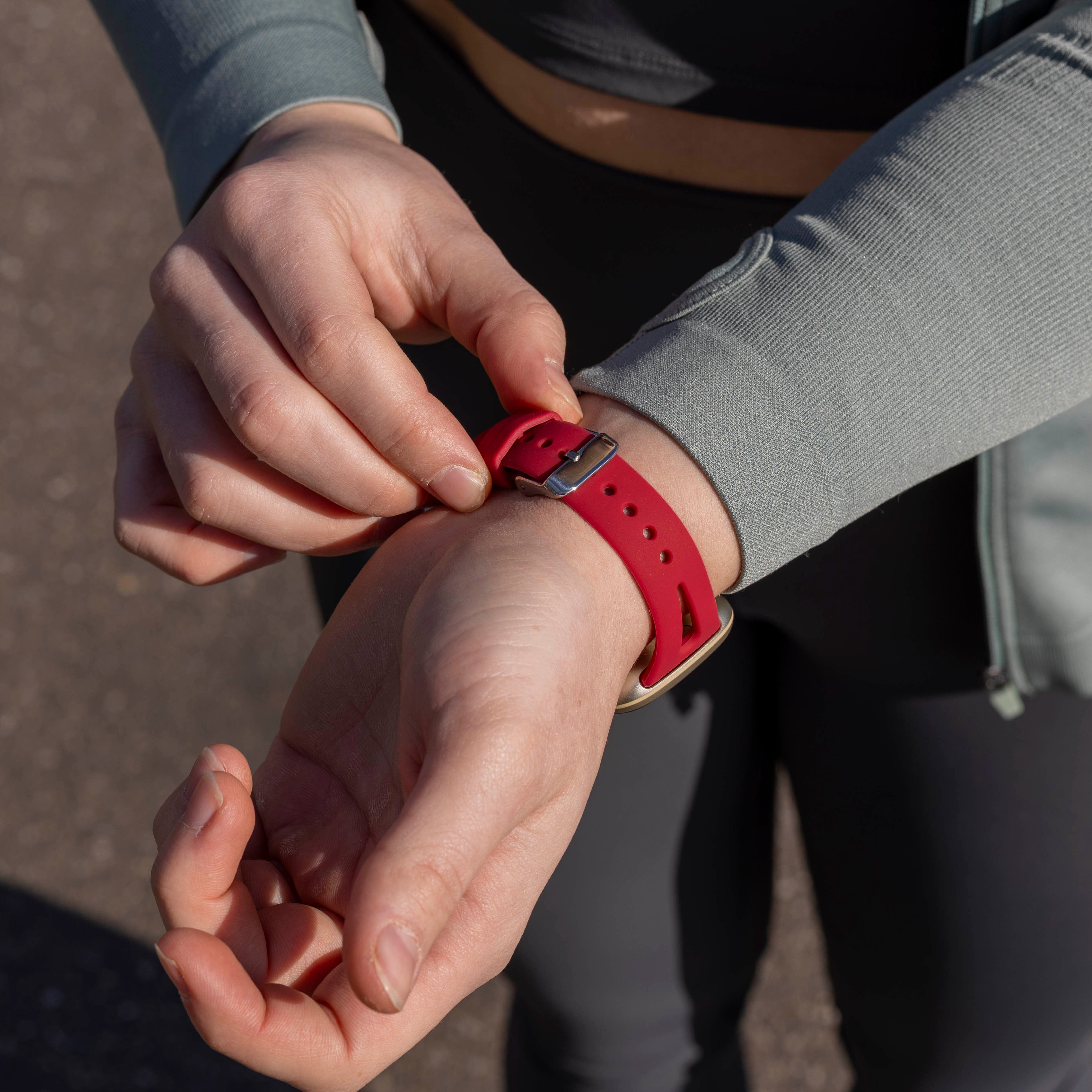 Eine Person stellt eine rote Armbanduhr ein. Sie trägt eine graue Jacke und dunkle Kleidung. Der Hintergrund ist unscharf.
