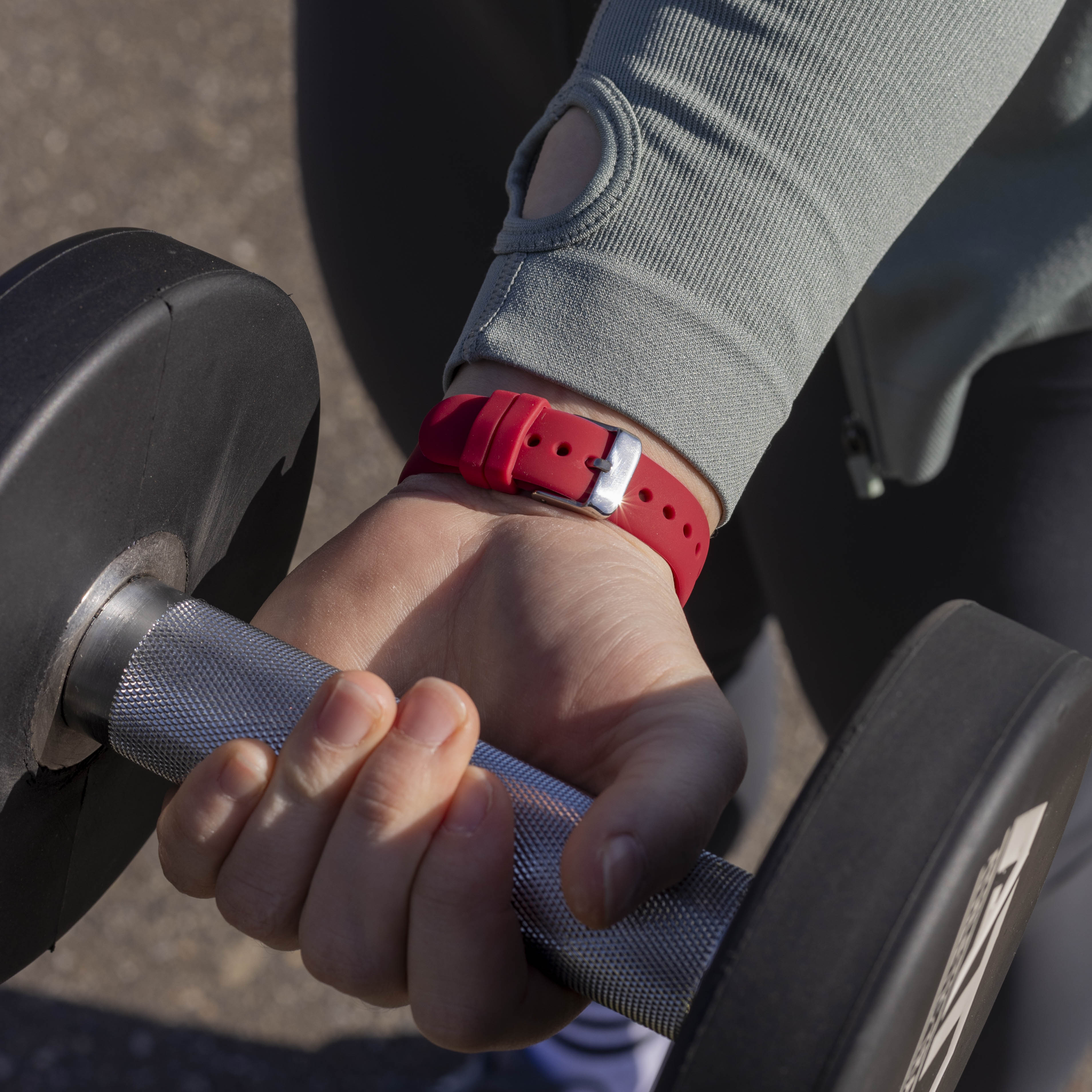 Eine Person hält eine Hantel. Am Handgelenk befindet sich ein rotes Armband. Die Person trägt ein graues Langarmshirt.