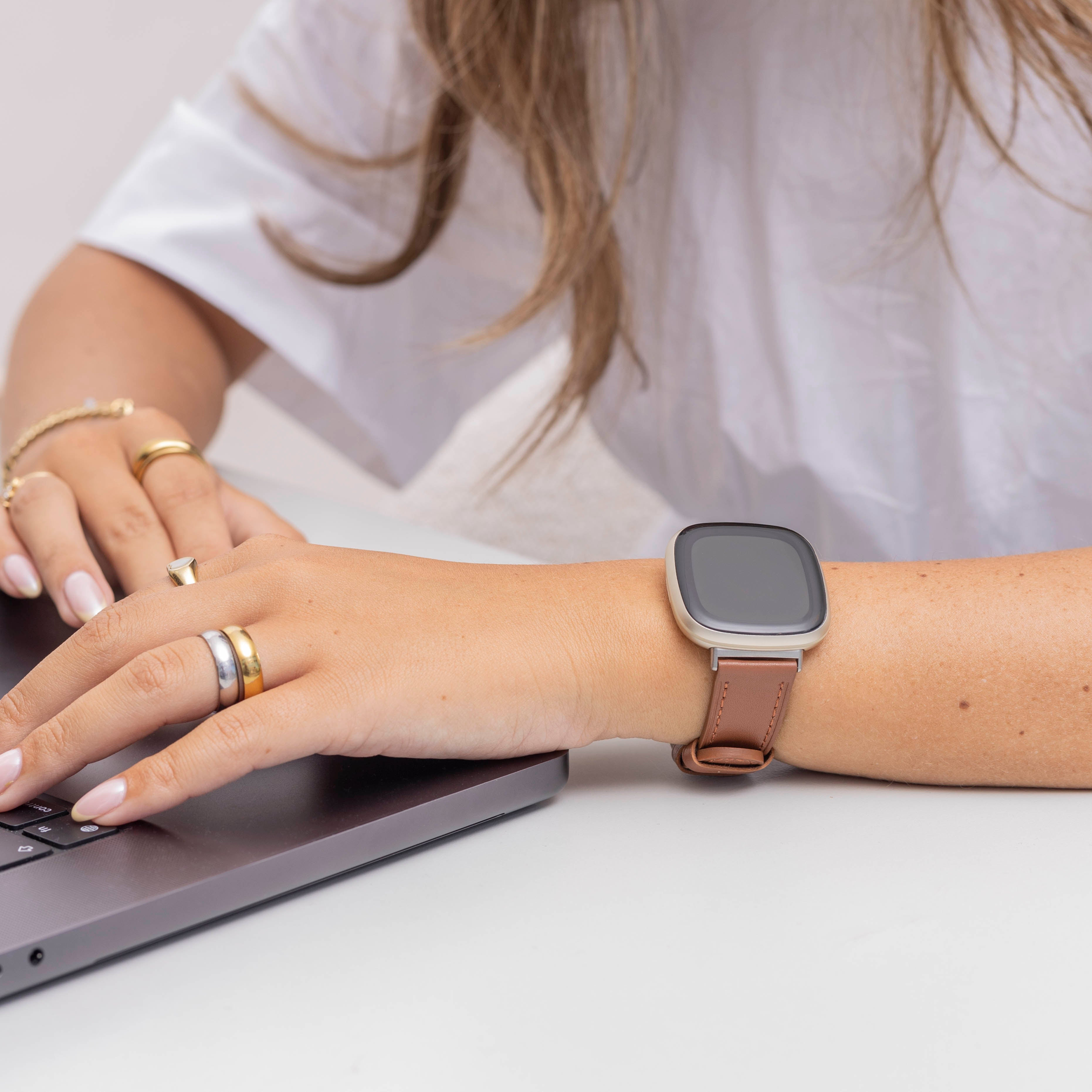 Frau benutzt Laptop, trägt Smartwatch mit braunem Lederarmband. Ringe an den Fingern. Weißes Hemd.