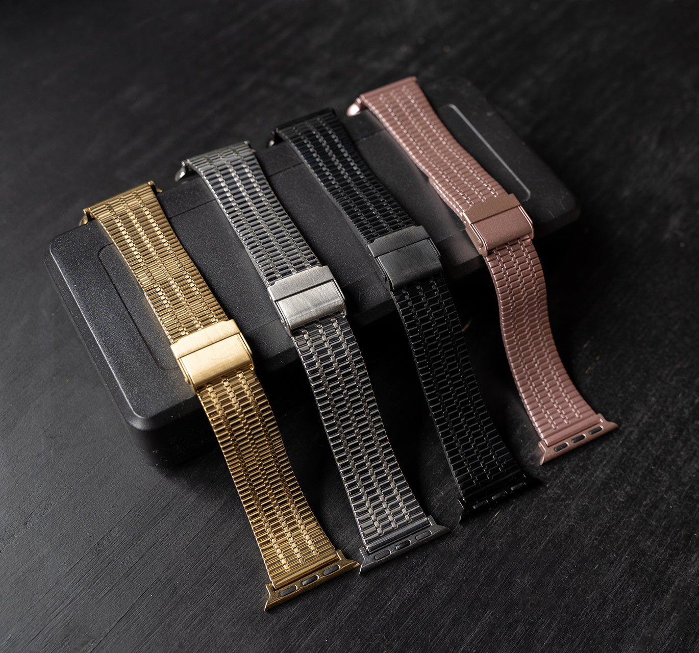 Fünf Uhrenarmbänder in Gold, Silber, Schwarz und Roségold. Sie liegen auf einer schwarzen Box auf dunkler Oberfläche.