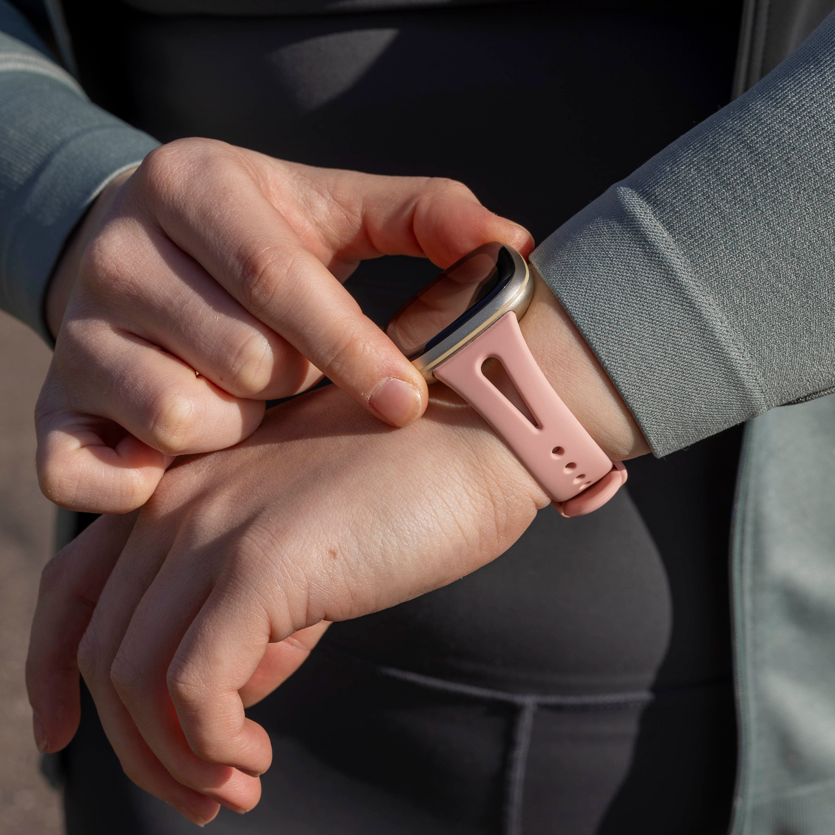 Hände einer Person, die eine Smartwatch mit rosa Armband einstellen. Die Person trägt eine graue Jacke und schwarze Sportkleidung.