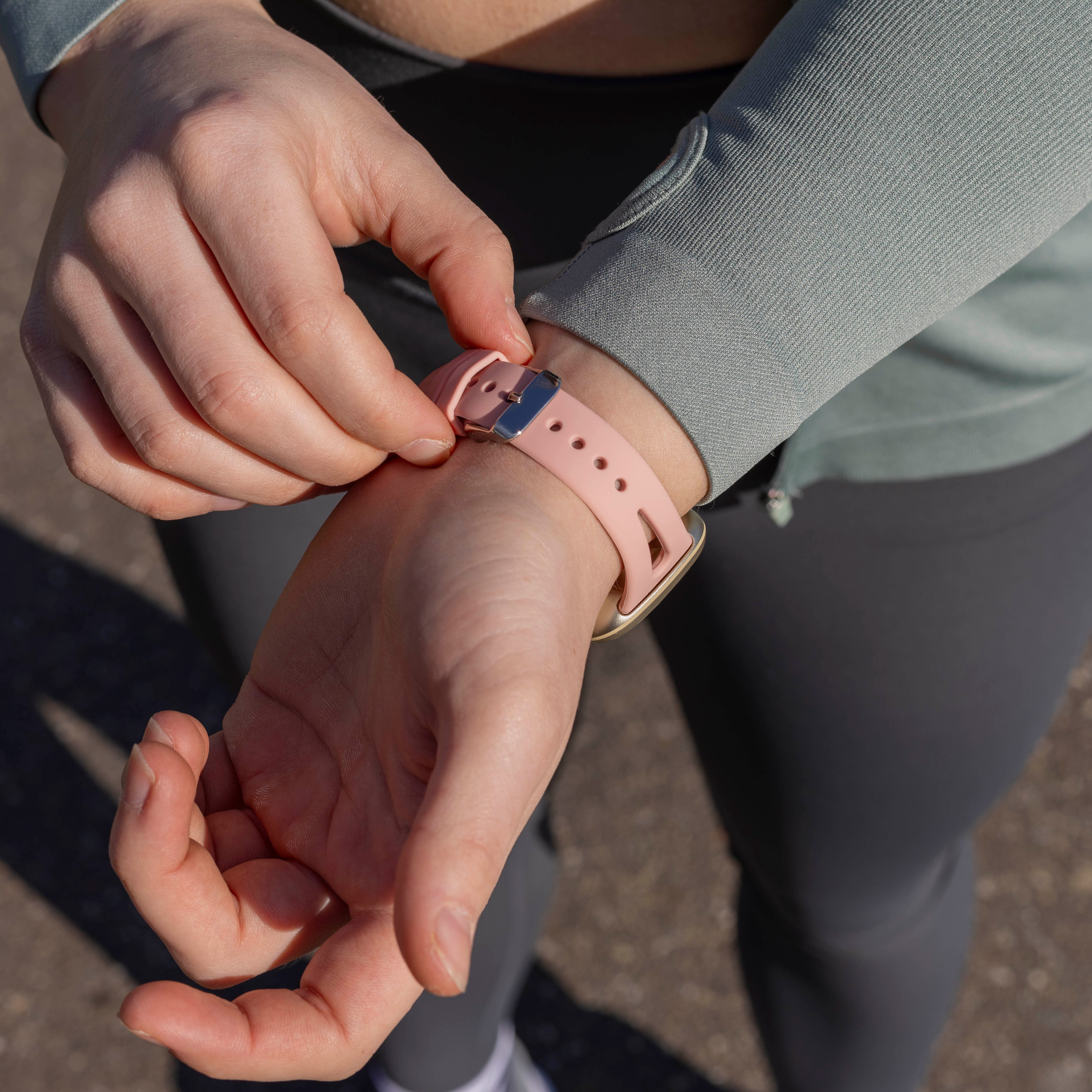 Hände einer Person, die eine rosa Smartwatch einstellt. Sie trägt graue Leggings und ein grünes Oberteil. Der Hintergrund ist unscharf.