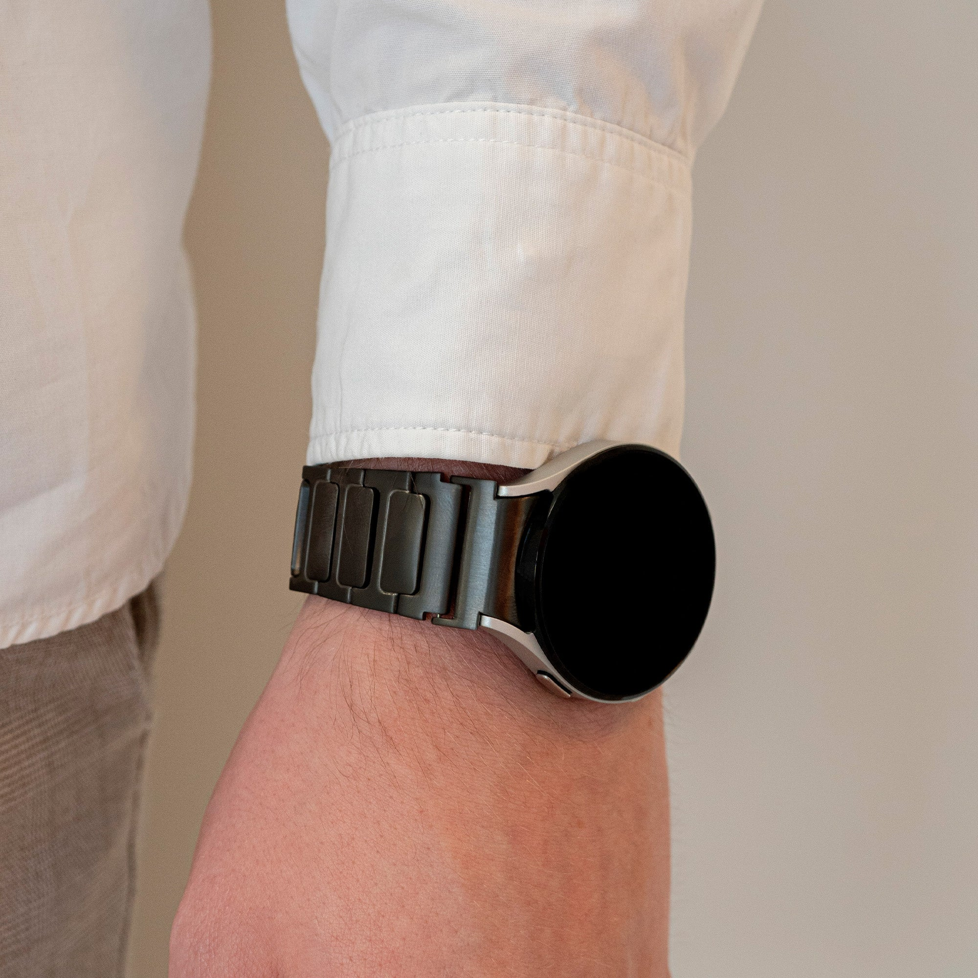 Nahaufnahme eines Handgelenks mit einer schwarzen Smartwatch mit Metallarmband, weißem Hemdärmel und hellbrauner Hose.
