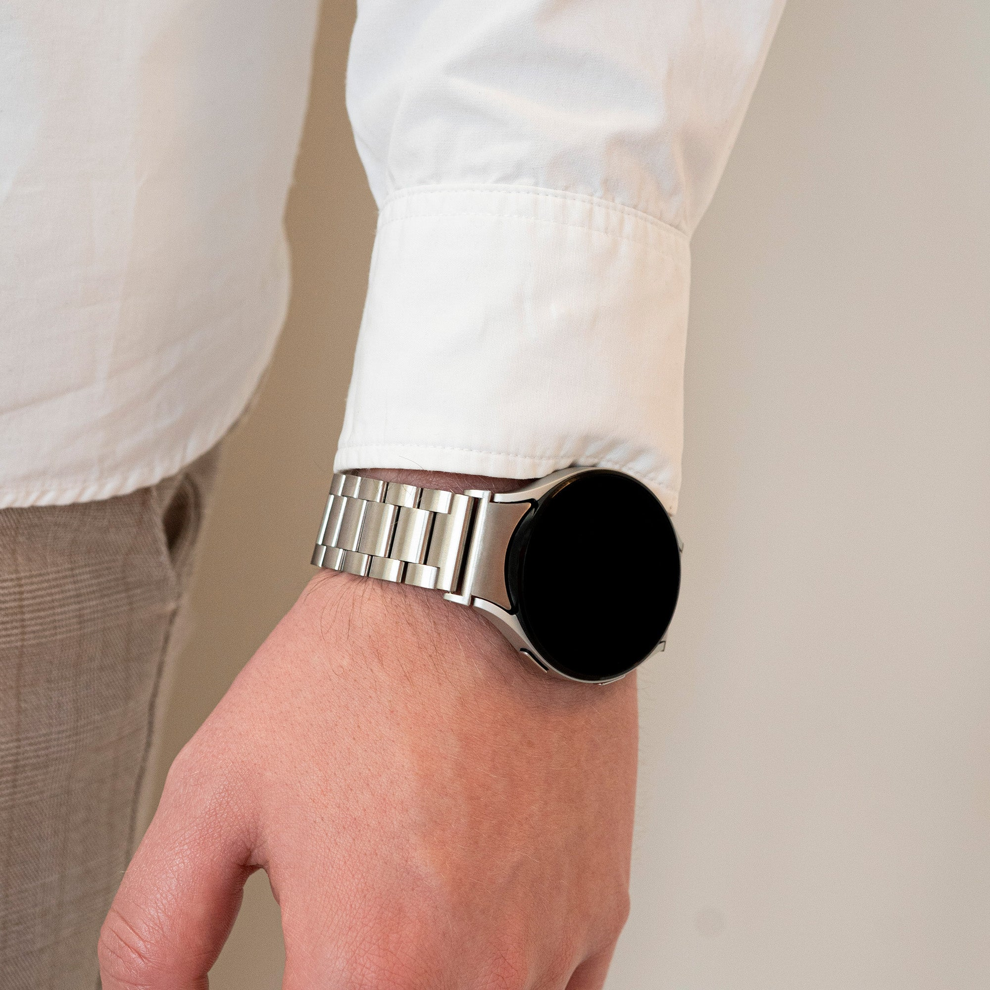 Handgelenk einer Person mit silberner Smartwatch. Die Person trägt ein weißes Hemd und beige Hose. Der Hintergrund ist cremefarben.