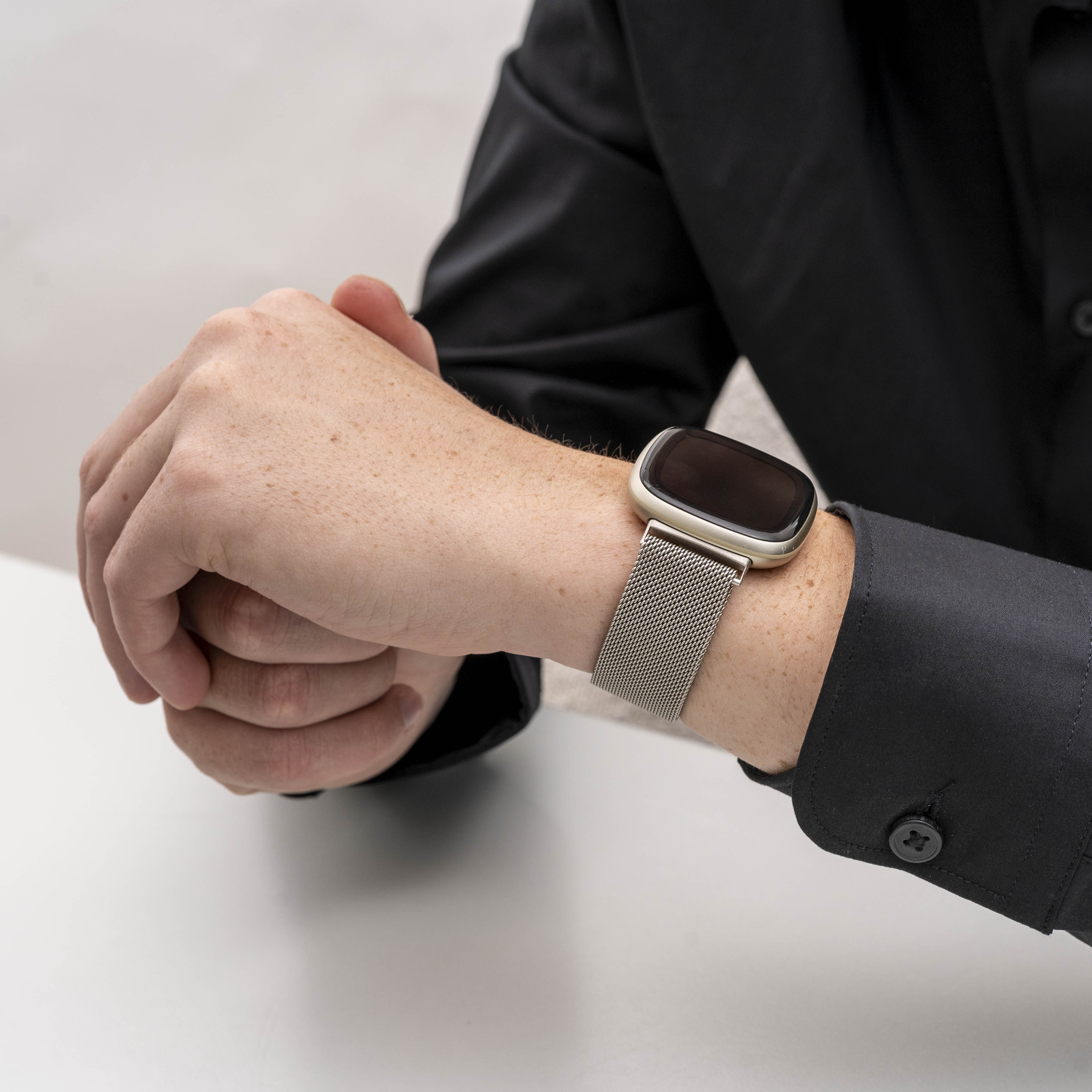 Handgelenk mit Smartwatch, schwarzes Hemd. Hände gefaltet, Uhr hat silbernes Mesh-Armband und helles Ziffernblatt.