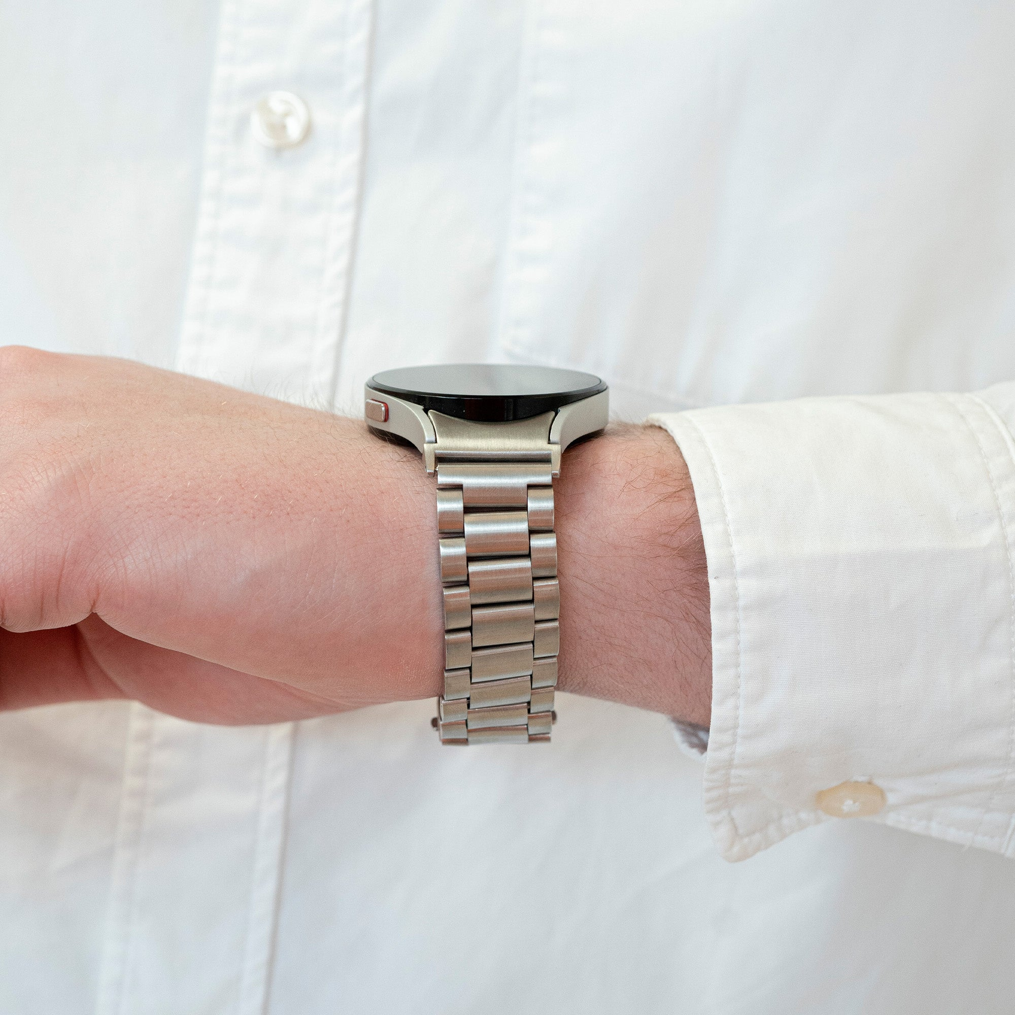 Das Handgelenk einer Person mit einer silbernen Smartwatch. Die Uhr ist metallisch. Der Hintergrund ist ein weißes Hemd.