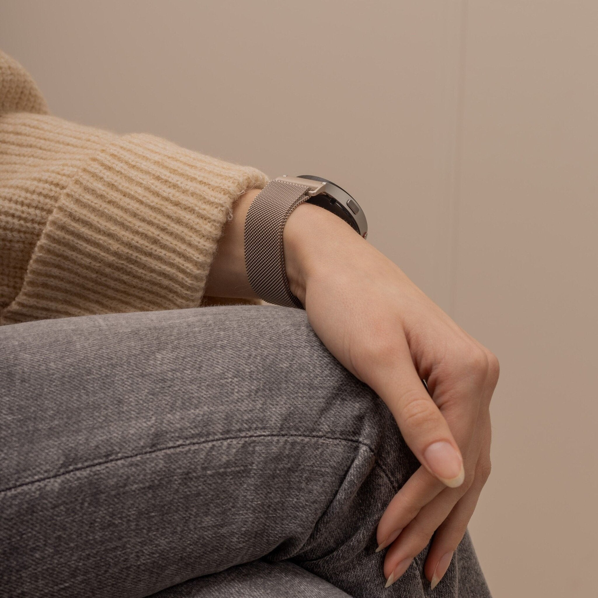 Ein Arm mit Uhr am Handgelenk und Hand, die auf Jeans ruht. Beiger Pullover, graue Jeans und ein neutraler Hintergrund.