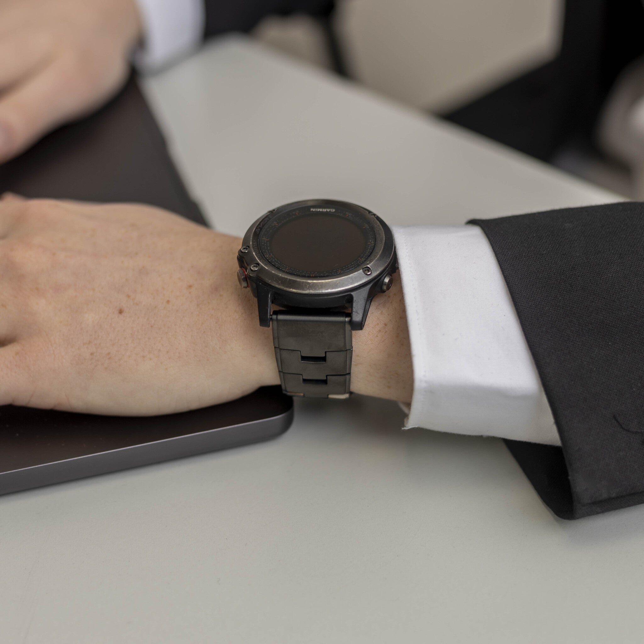 Das Handgelenk einer Person mit einer schwarzen Garmin-Uhr. Die Person trägt ein weißes Hemd und einen schwarzen Anzug. Ein Laptop ist sichtbar.