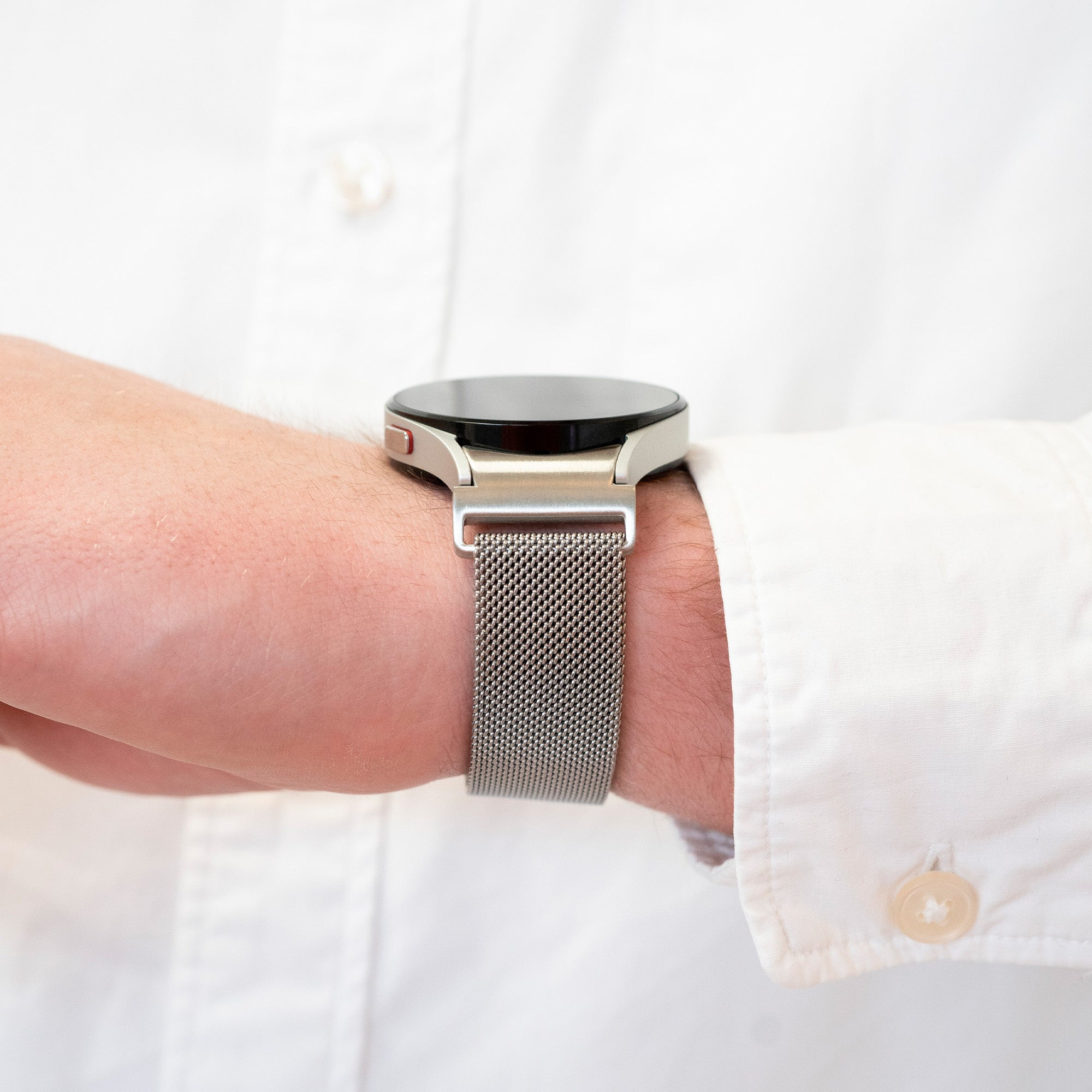 Eine silberne Smartwatch an einem Handgelenk mit Mesh-Armband. Die Person trägt ein weißes Hemd.