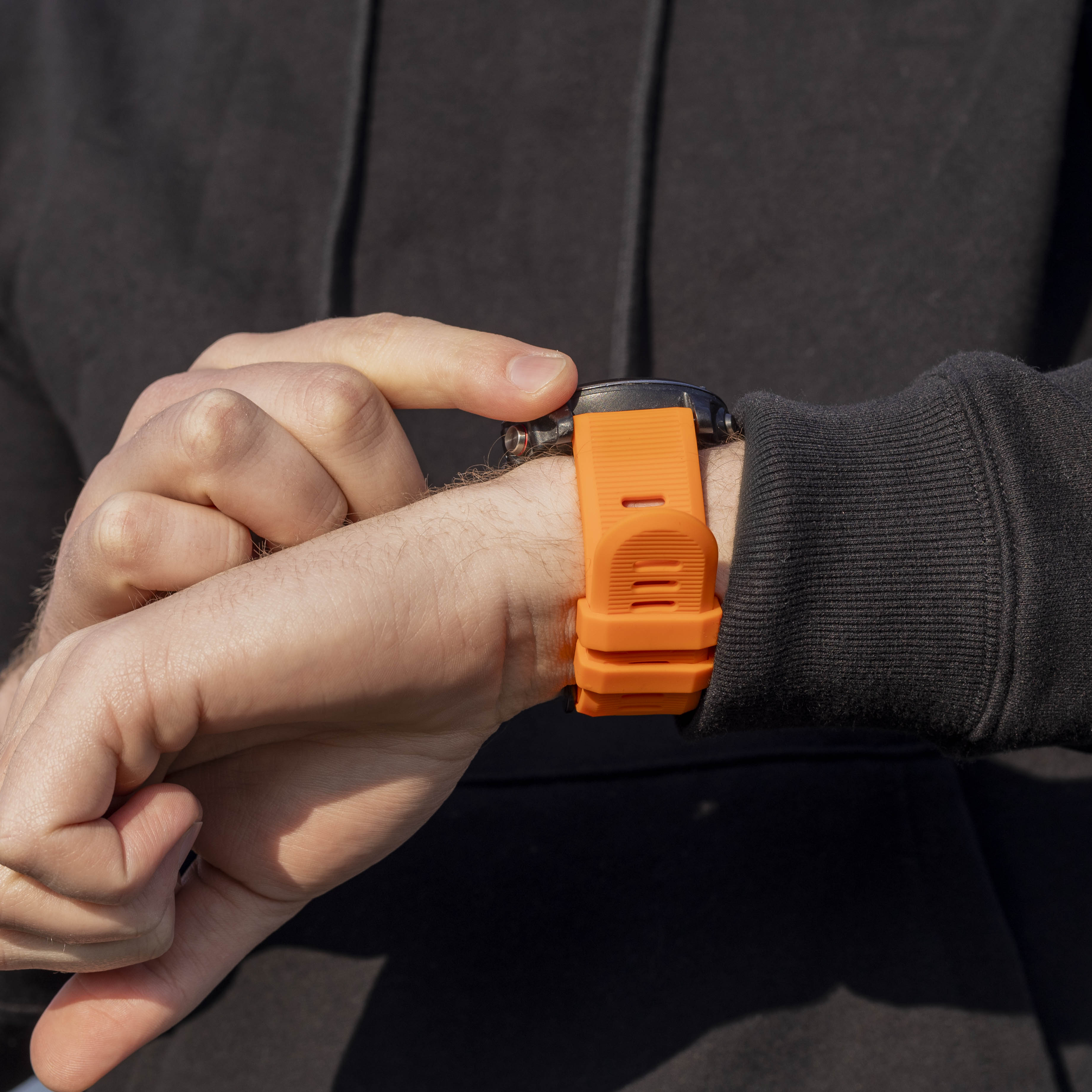 Die Hand einer Person mit einer orangefarbenen Smartwatch. Der Finger der Person berührt die Uhr. Dunkler Hintergrund und schwarzer Kapuzenpullover.