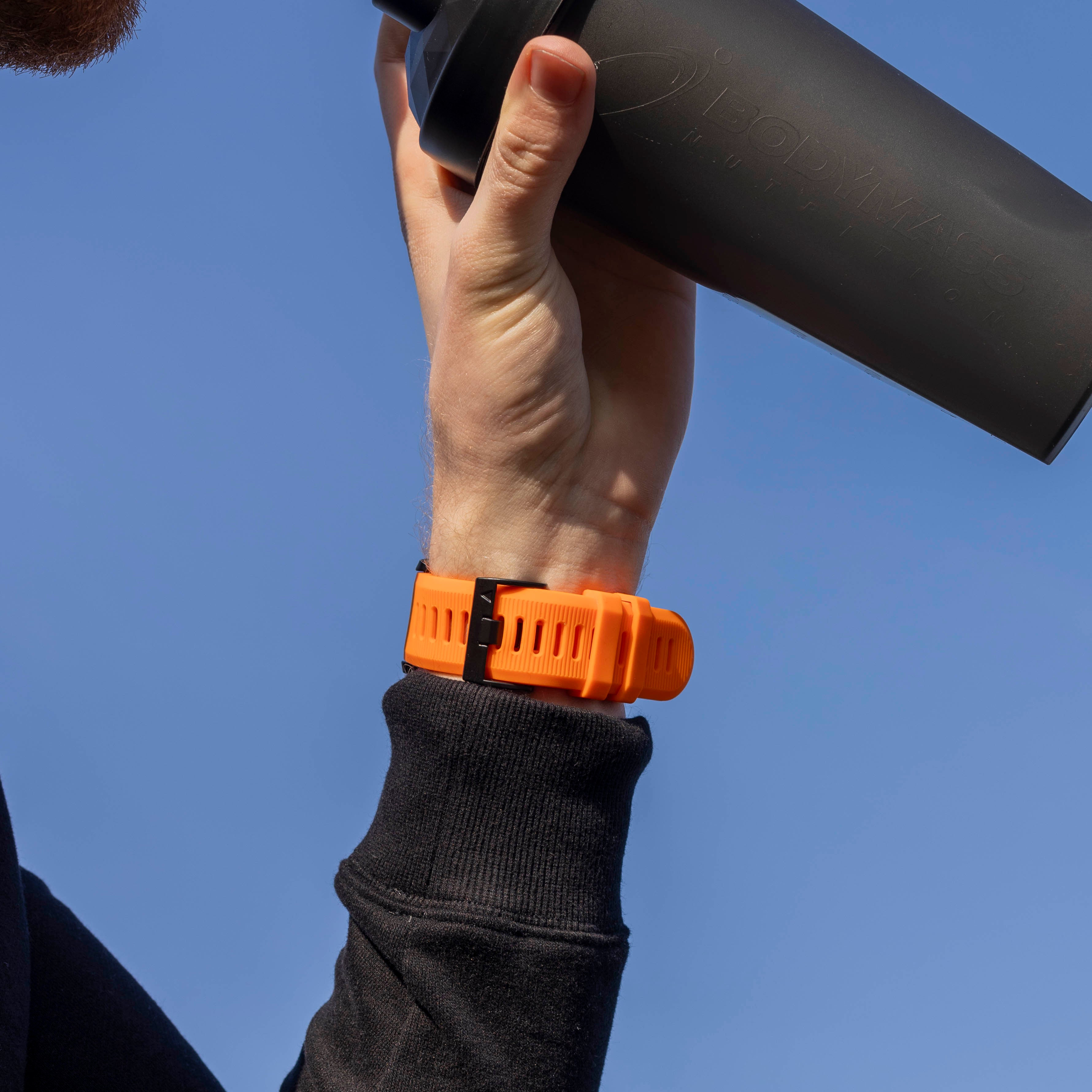 Eine Person, die einen schwarzen Shaker hält, mit einer orangefarbenen Uhr am Handgelenk vor blauem Himmel.