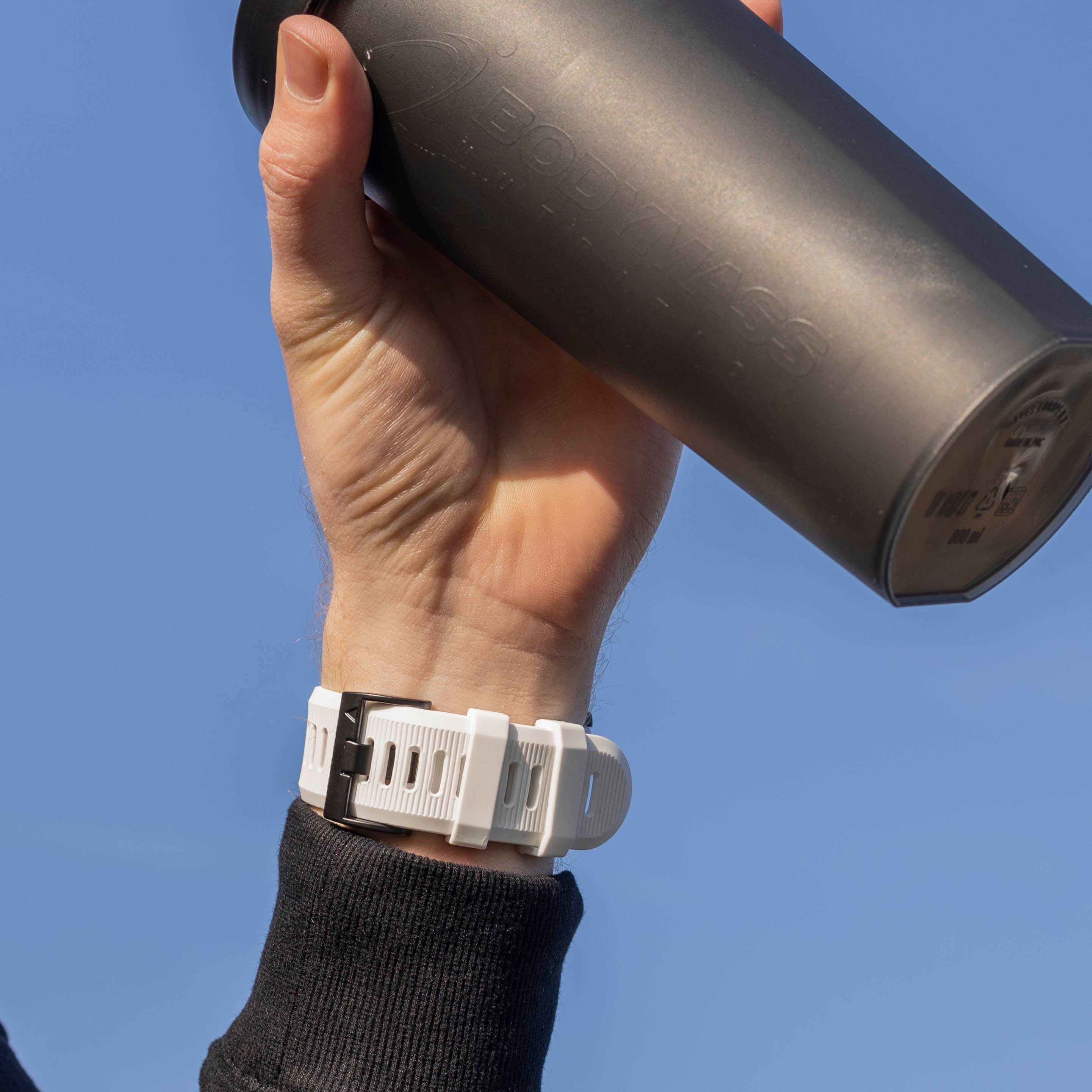 Eine Hand hält eine schwarze Wasserflasche mit einer weißen Uhr. Der Hintergrund ist ein klarer blauer Himmel.