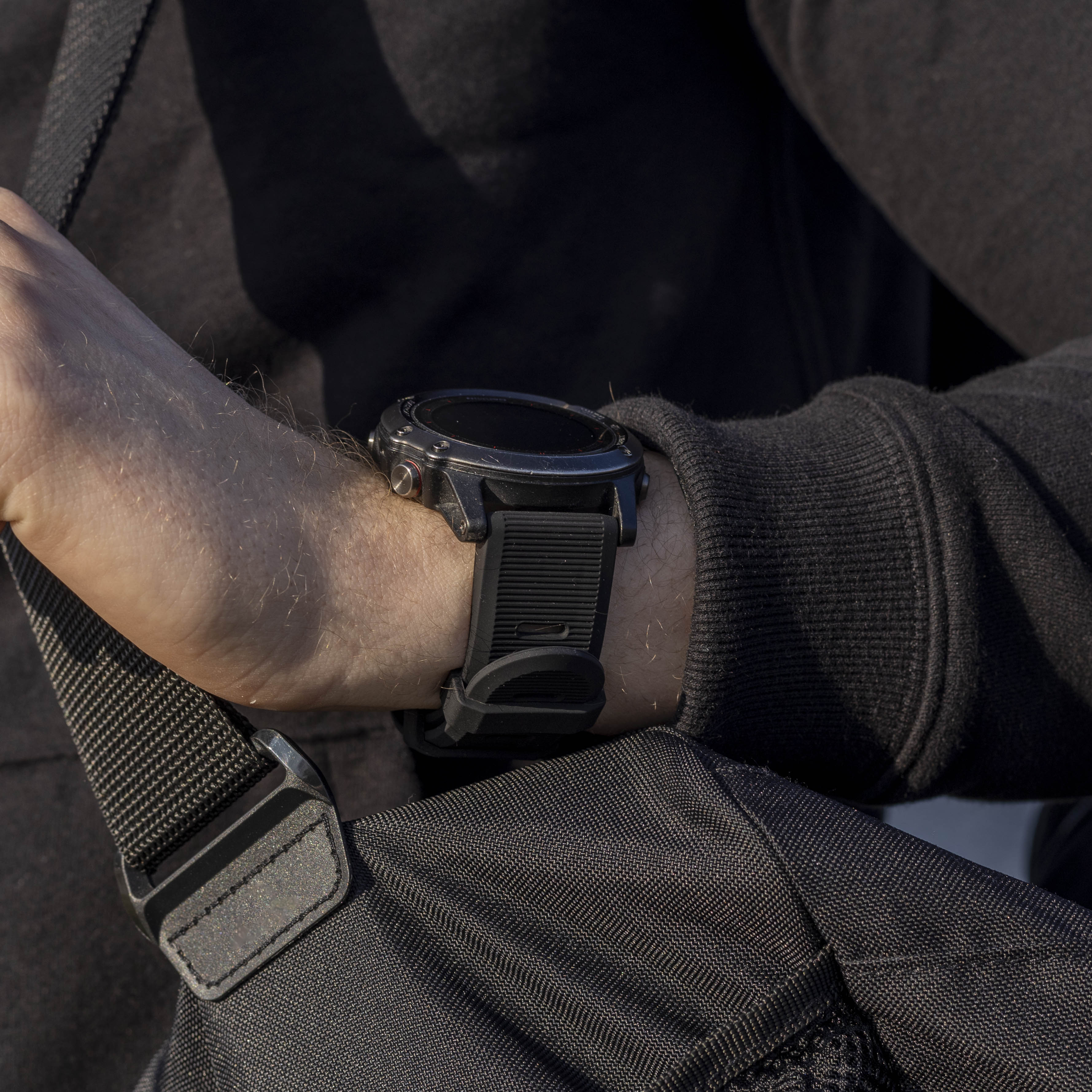 Der Arm einer Person mit einer schwarzen Smartwatch am Handgelenk. Schwarzes Armband. Schwarzer Pullover und eine schwarze Tasche sichtbar.