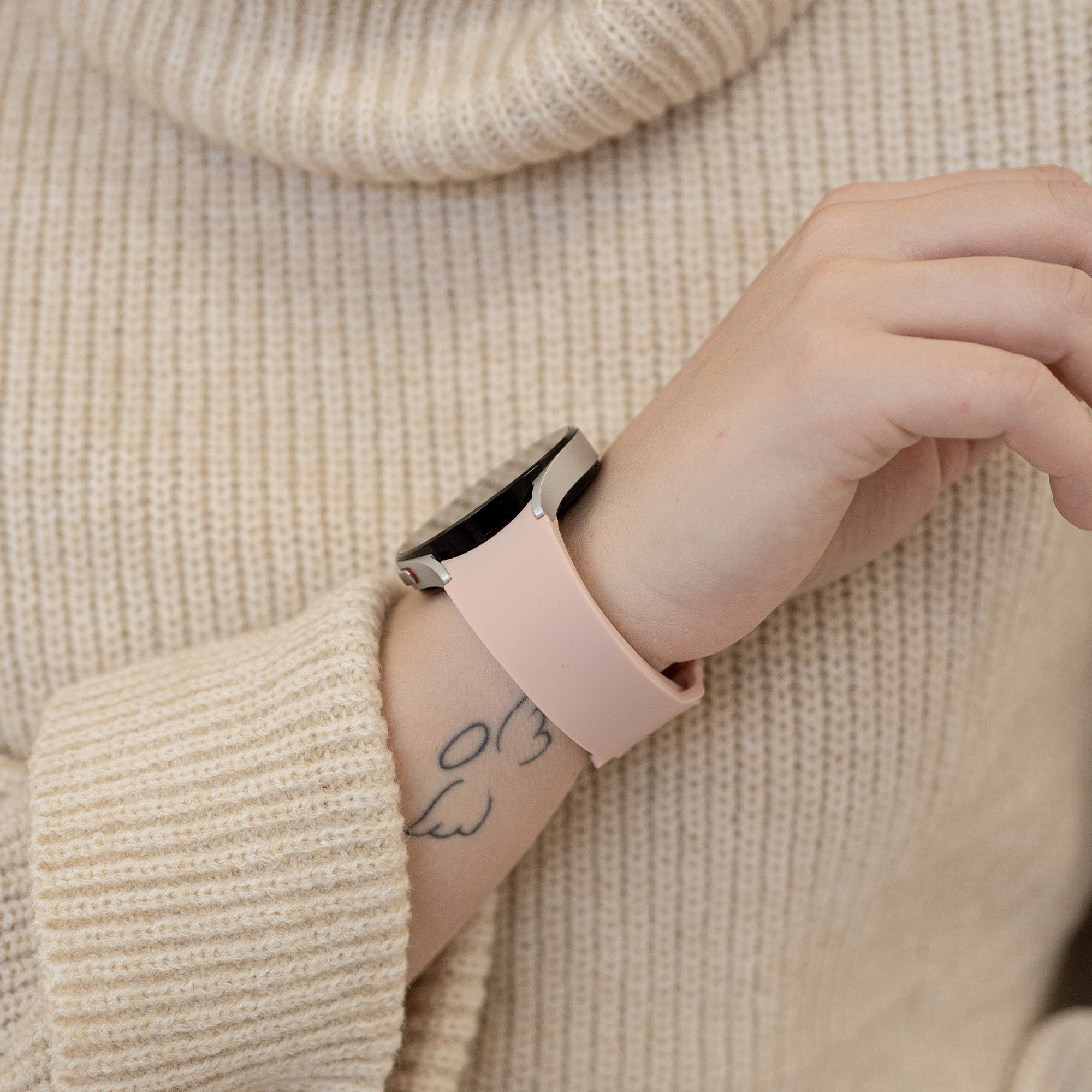 Das Handgelenk einer Person mit einer Smartwatch. Die Uhr hat ein rosa Armband. Die Person trägt einen cremefarbenen Pullover.