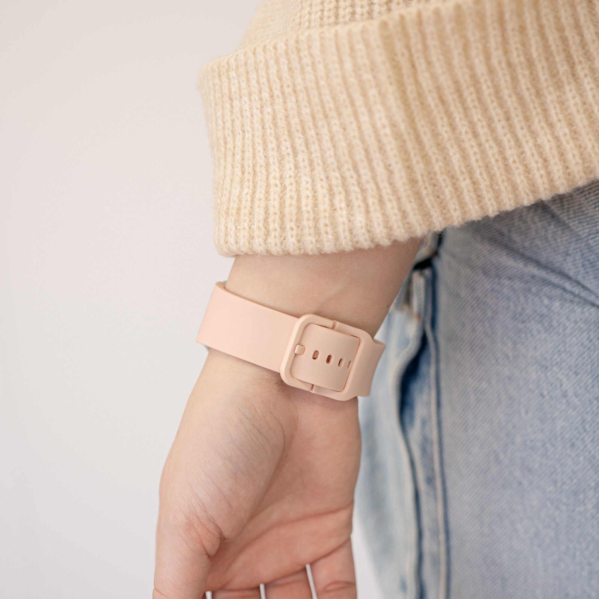 Nahaufnahme eines Handgelenks mit einer hellrosa Uhr. Beige Pullover und blaue Jeans sind ebenfalls sichtbar.