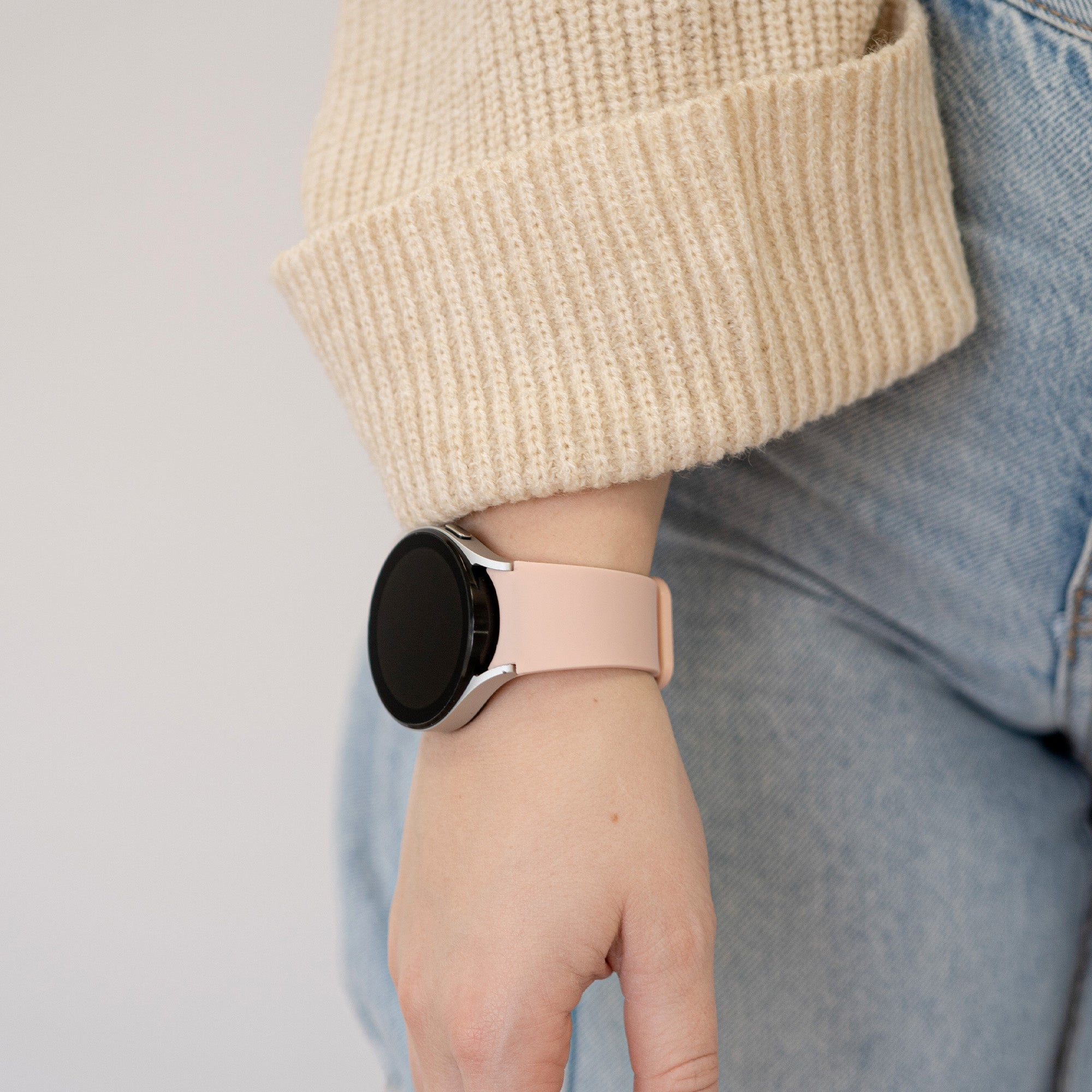 Eine Person, die eine Smartwatch mit rosa Armband trägt. Sie trägt einen beigen Pullover und blaue Jeans.