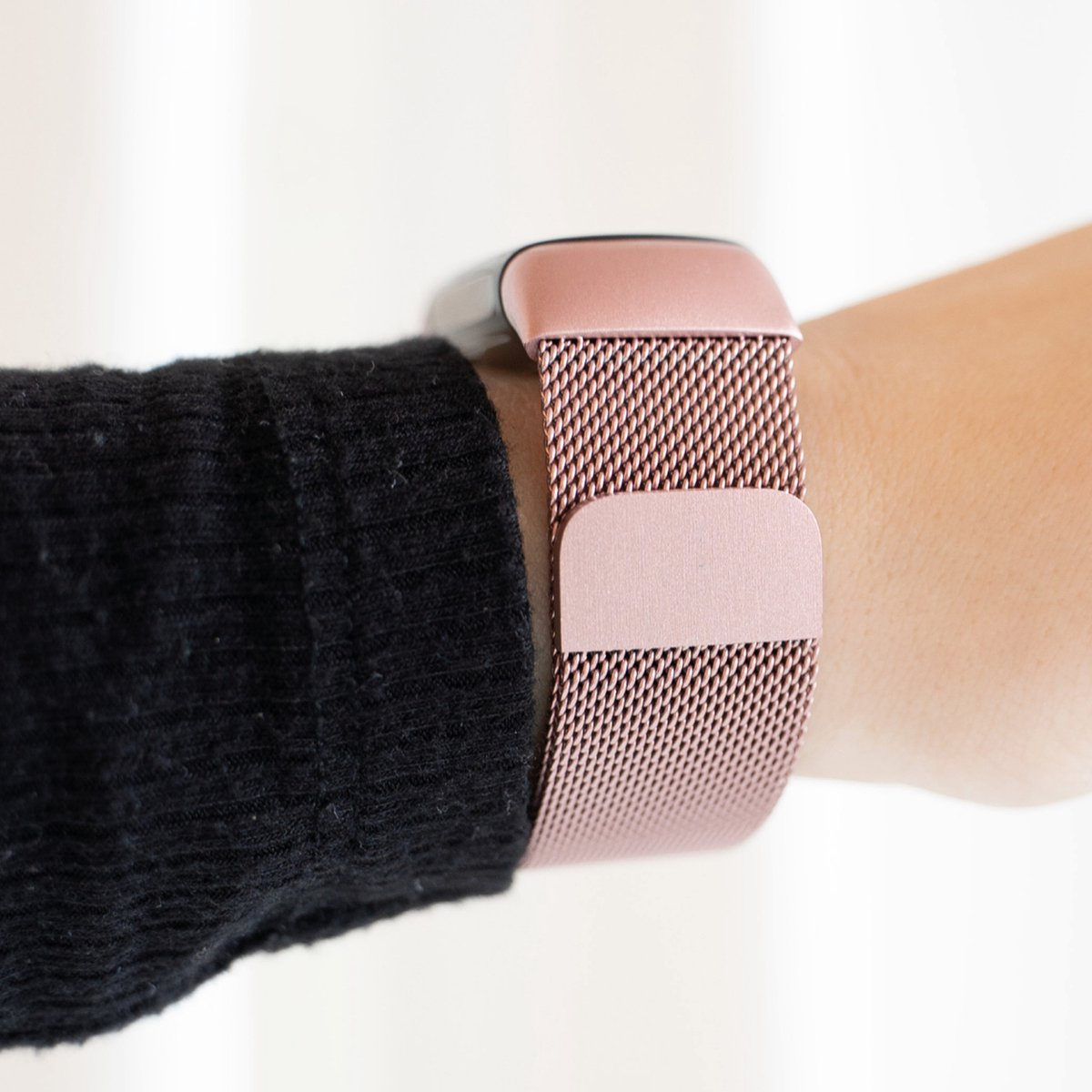 Ein Handgelenk mit einer roségoldenen Uhr. Das Armband ist aus Mesh. Der Ärmel ist schwarz. Der Hintergrund ist weiß.
