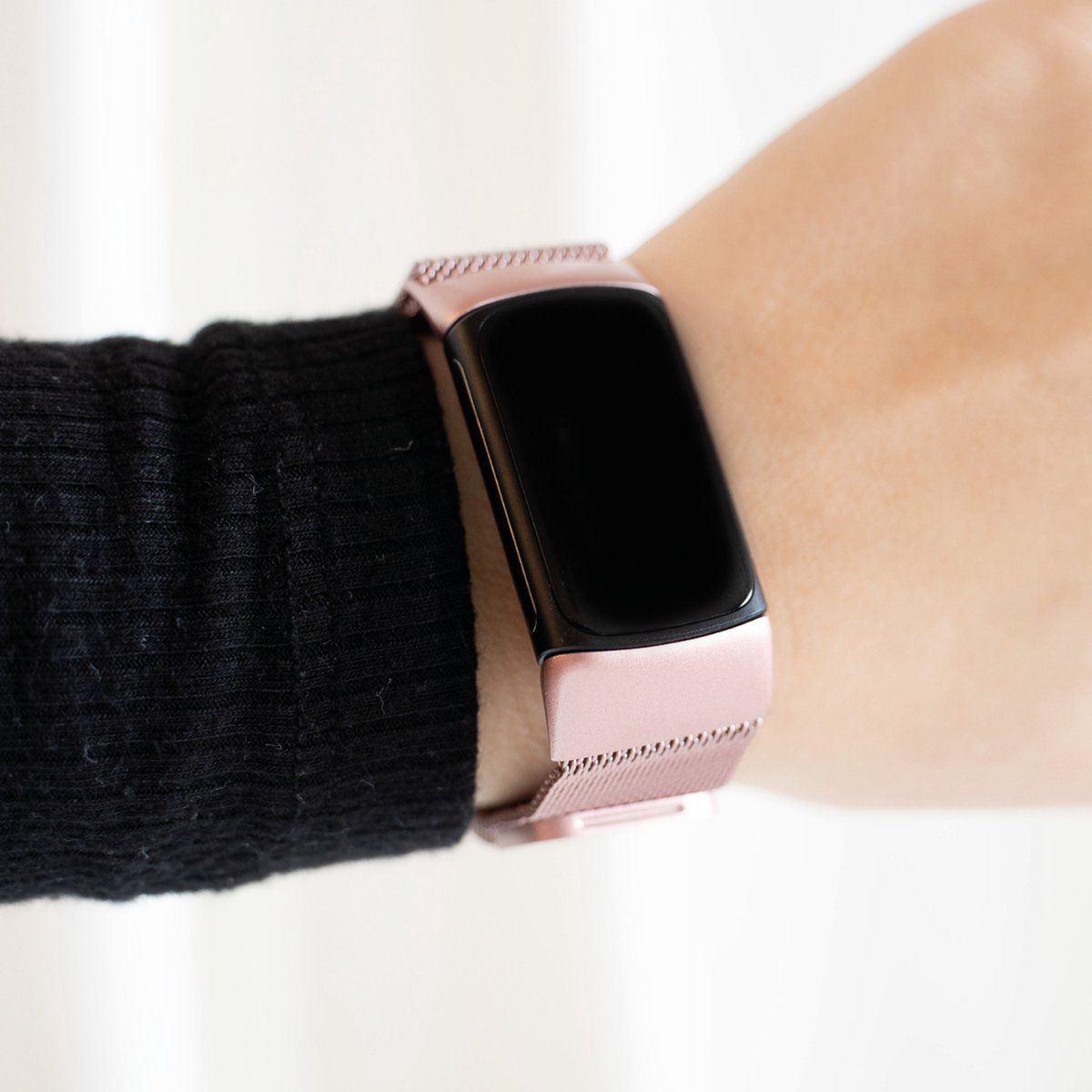 Handgelenk mit roségoldener Smartwatch. Die Uhr hat einen schwarzen Bildschirm und ein Mesh-Armband. Ein schwarzer Ärmel ist sichtbar.