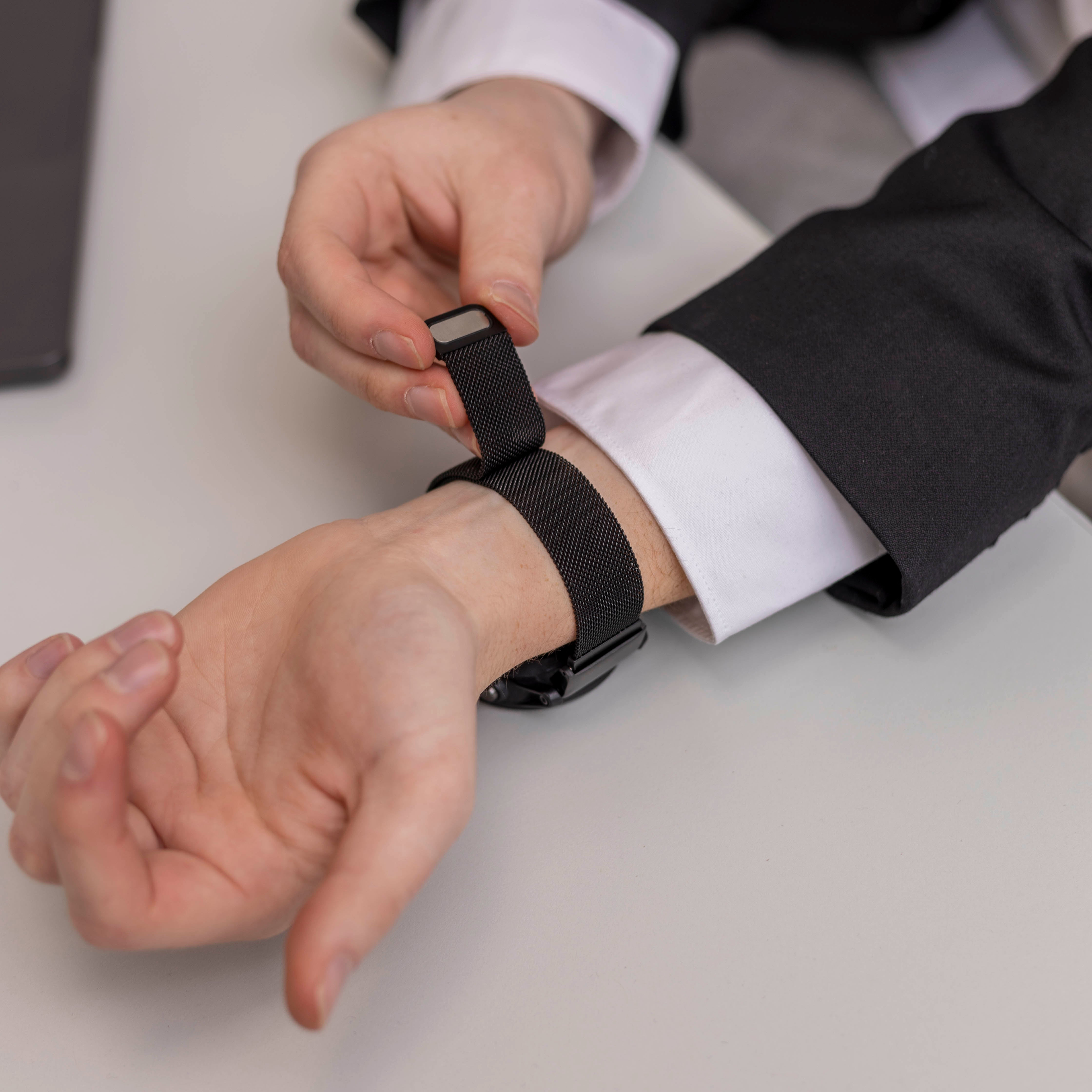 Eine Person stellt eine schwarze Uhr an ihrem Handgelenk ein. Sie trägt ein weißes Hemd und eine schwarze Anzugjacke. Der Hintergrund ist eine weiße Oberfläche.