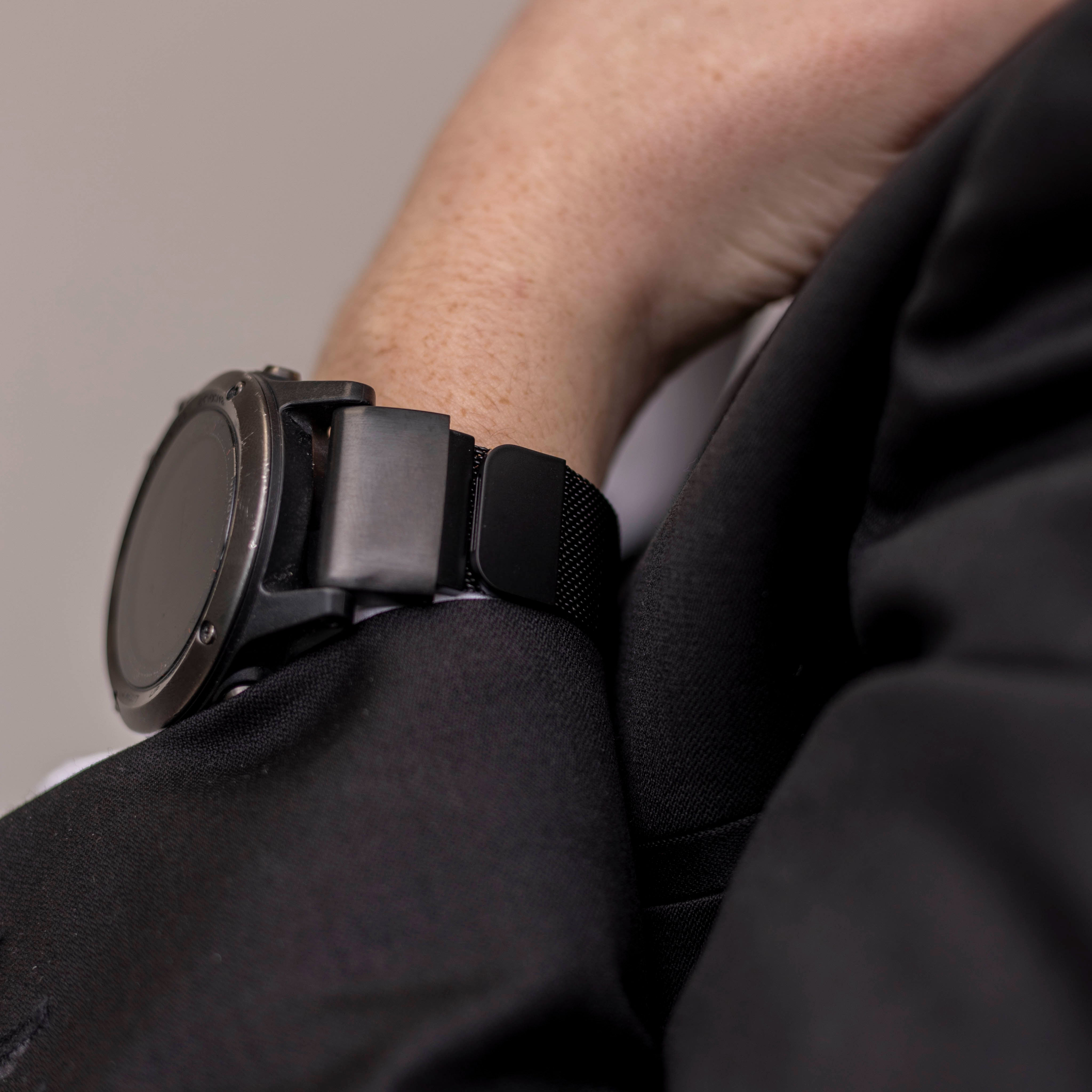 Eine schwarze Smartwatch befindet sich an einem Handgelenk. Das Handgelenk hat Sommersprossen und liegt neben einem schwarzen Blazer.
