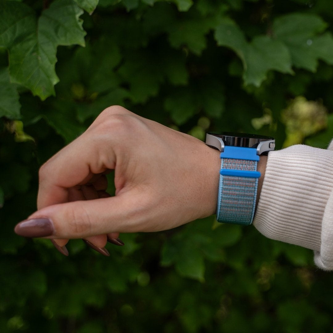 Eine Hand mit einer Smartwatch mit blauem und tealfarbenem Armband. Der Hintergrund zeigt grünes Laub. Die Person trägt einen cremefarbenen Pullover.