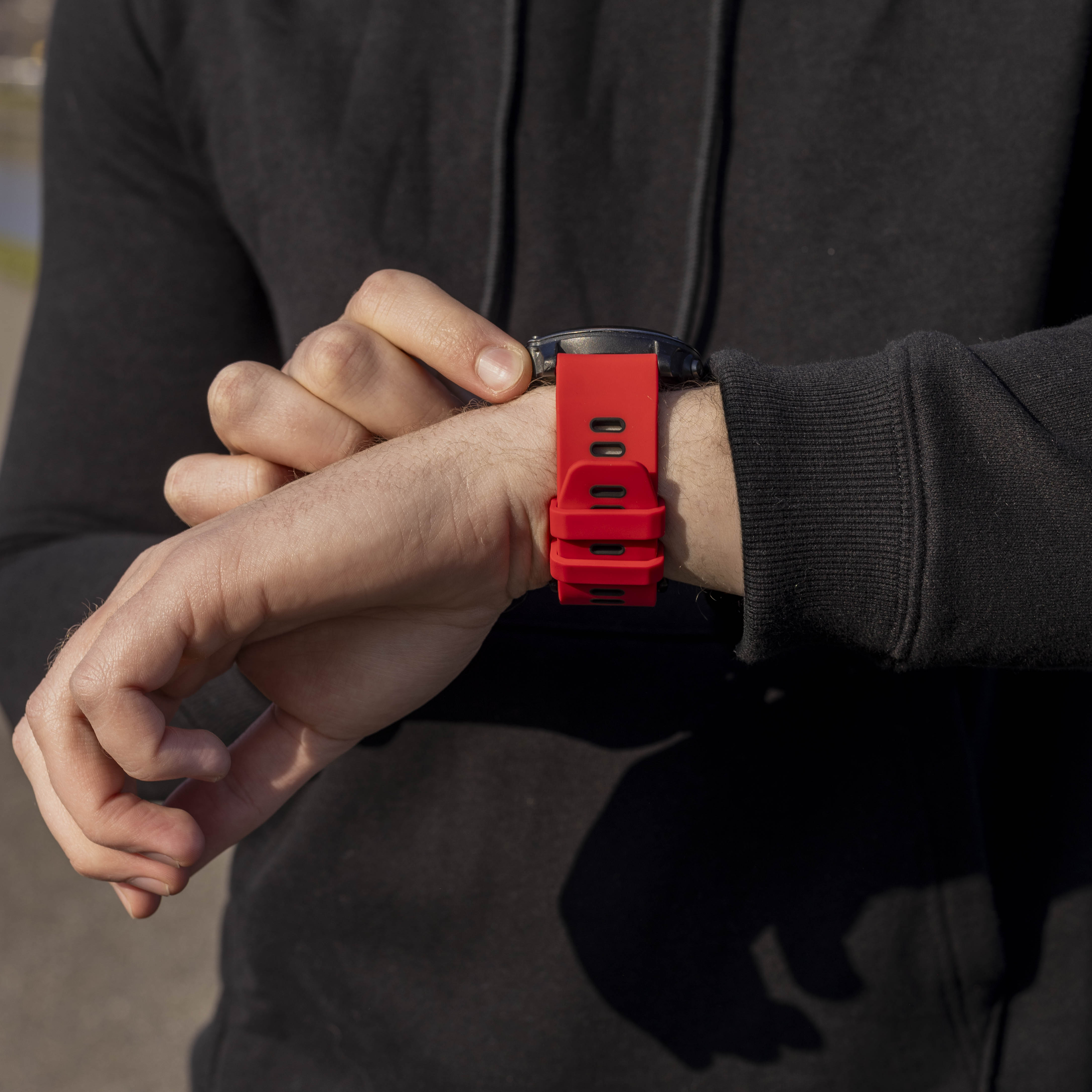 Person im schwarzen Kapuzenpullover überprüft eine Armbanduhr mit rotem Armband. Die Uhr ist schwarz, der Hintergrund unscharf.