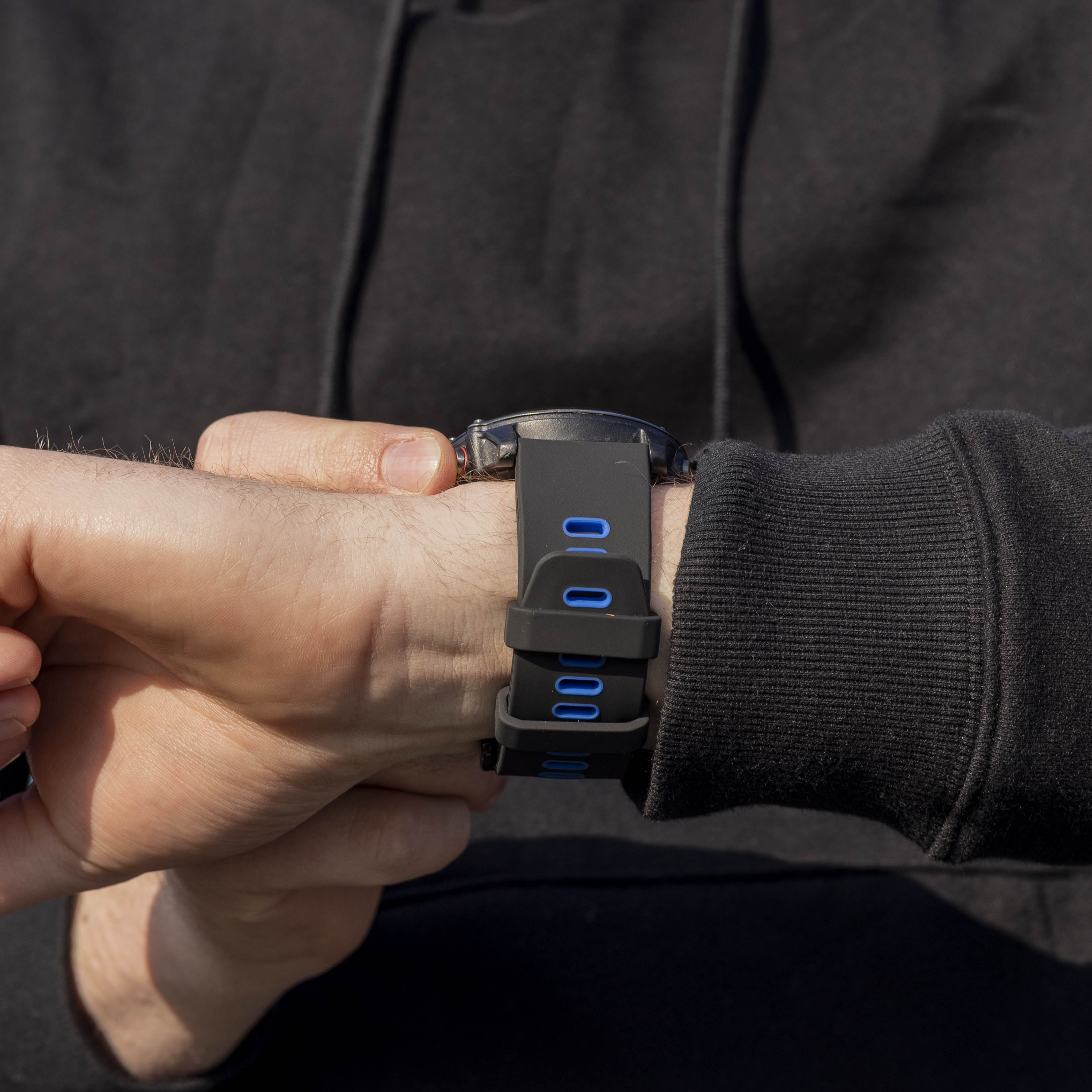 Handgelenk mit schwarzer Uhr, blauen Akzenten und einem schwarzen Kapuzenärmel im Hintergrund.