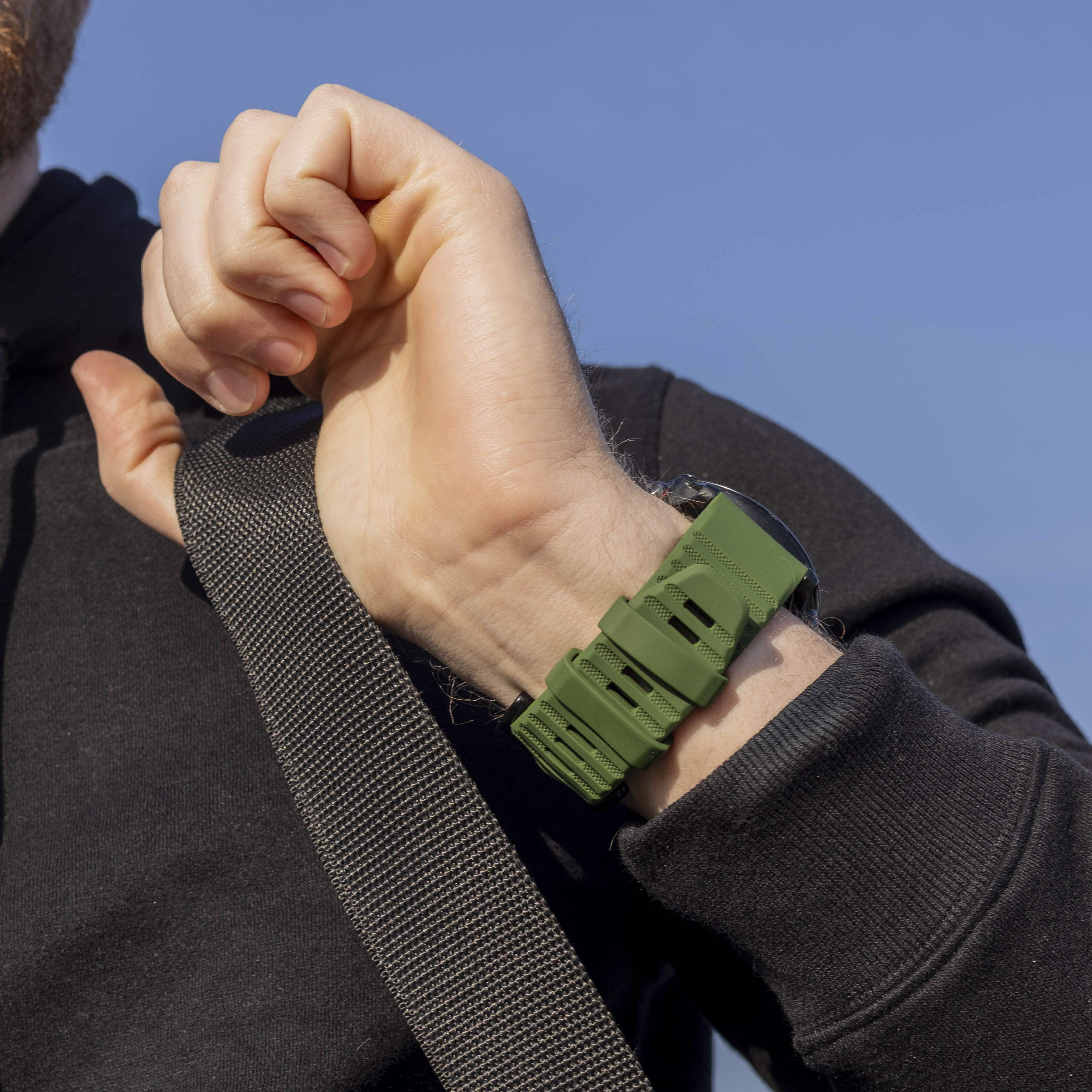 Ein Arm mit grüner Uhr und Armband. Person trägt schwarzen Hoodie, hält schwarzen Gurt vor blauem Himmel.