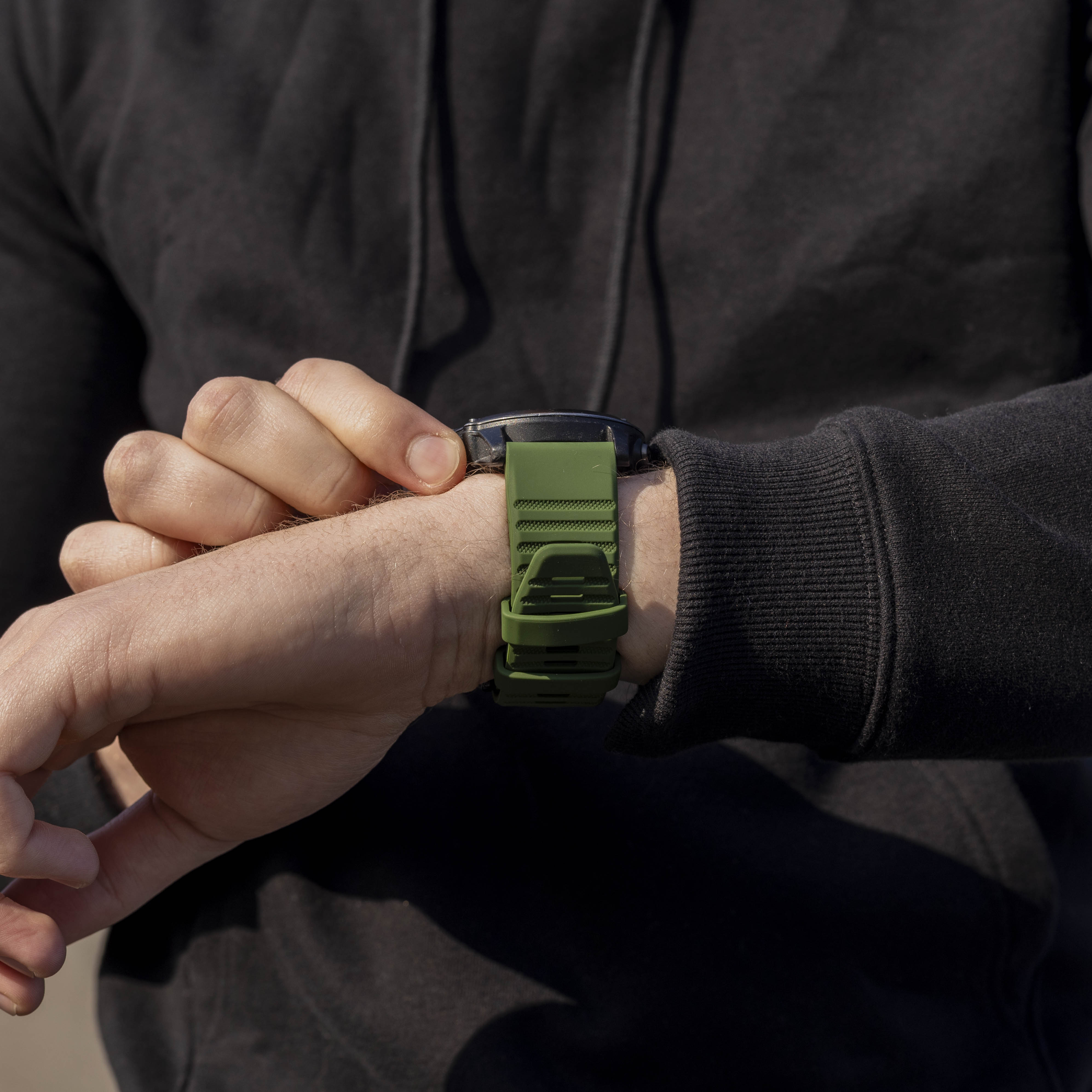 Eine Person im schwarzen Hoodie schaut auf ihre Handgelenk mit einer grünen Armbanduhr. Die Uhr ist schwarz, das Band olivgrün.