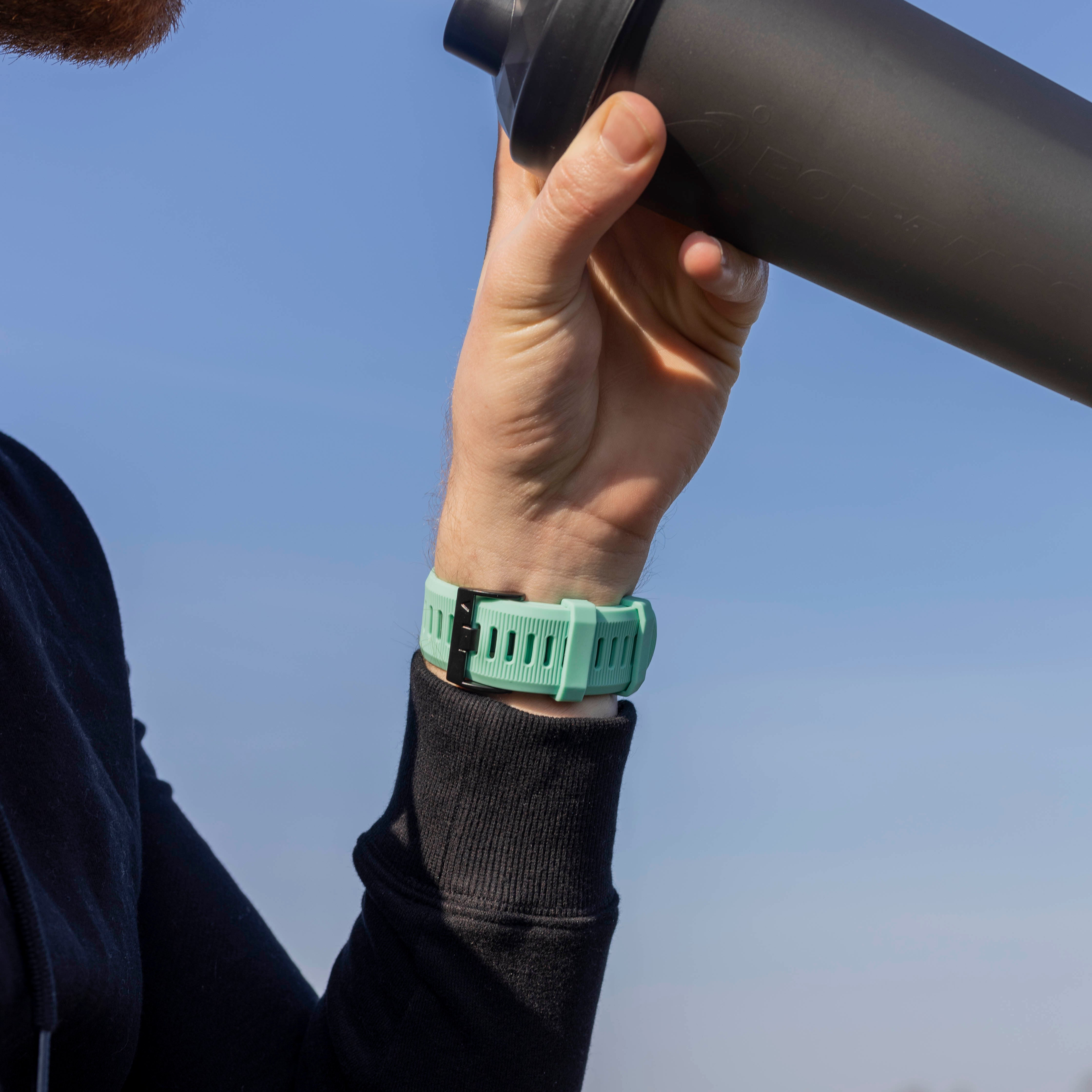 Eine Person hält eine schwarze Flasche und trägt eine mintfarbene Uhr. Der Hintergrund ist ein klarer blauer Himmel.