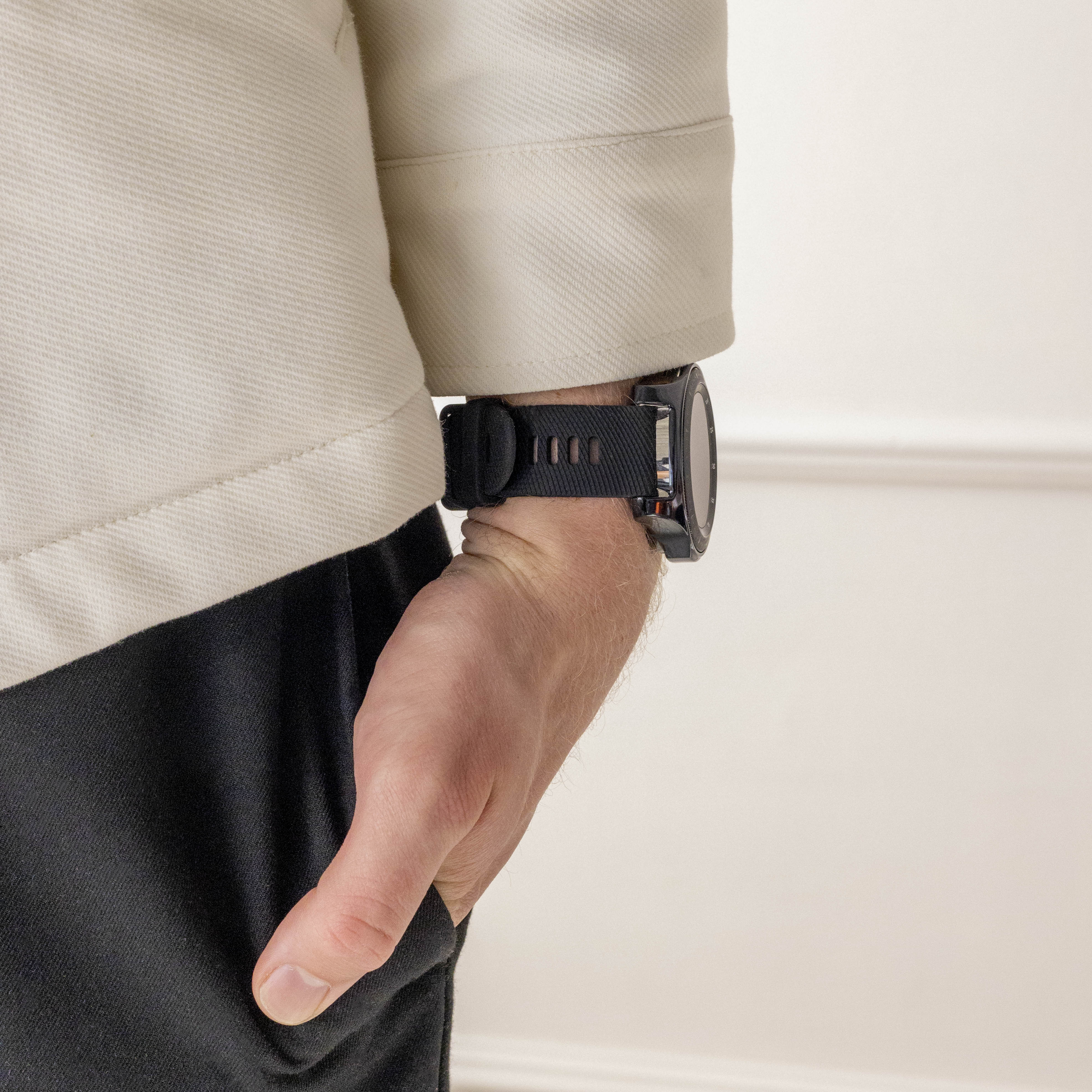 Ein Arm mit einer schwarzen Uhr ist zu sehen. Die Person trägt eine beige und schwarze Jacke. Der Hintergrund ist eine weiße Wand.