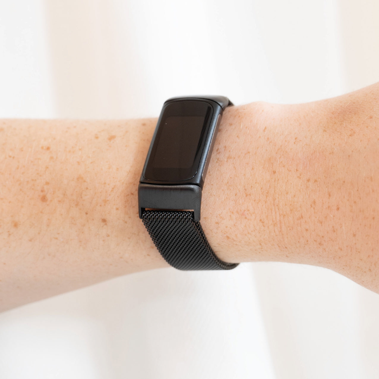Eine schwarze Smartwatch am Handgelenk. Die Uhr hat einen rechteckigen Bildschirm und ein Mesh-Armband. Die Haut hat Sommersprossen, und der Hintergrund ist weiß.