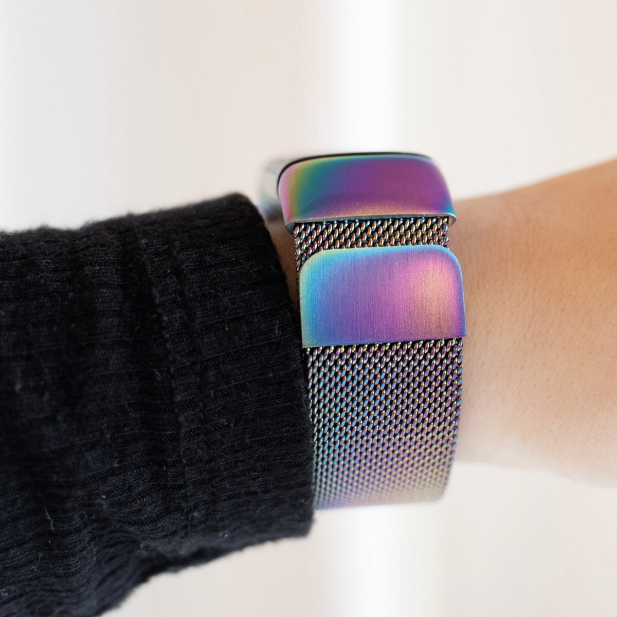 Ein Handgelenk mit einer regenbogenfarbenen Uhr. Das Uhrenarmband ist aus Mesh. Im Vordergrund ein schwarzer Ärmel und im Hintergrund ein weißer Hintergrund.