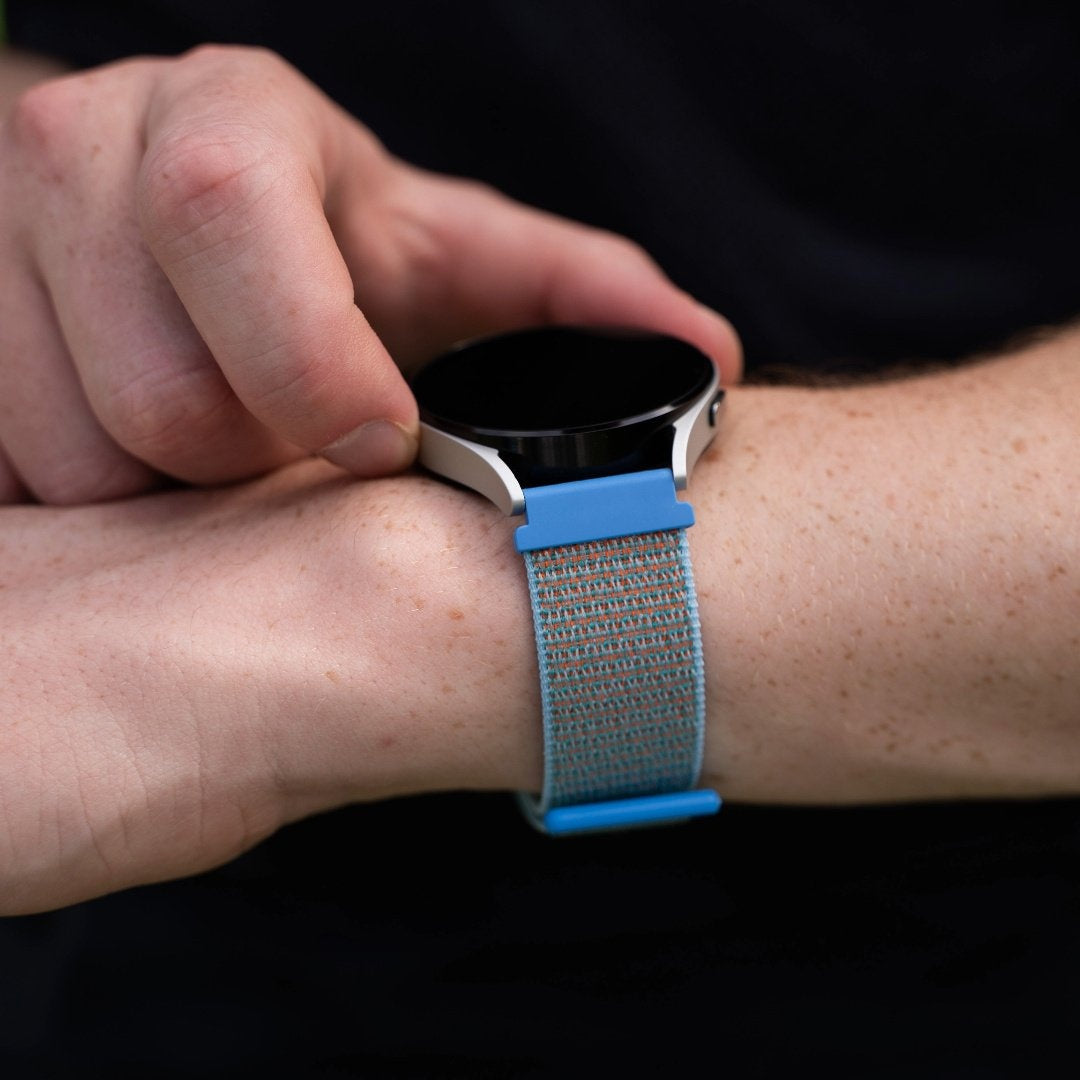 Ein Handgelenk mit Smartwatch. Die Uhr hat ein blaues Armband, ein silbernes Gehäuse und einen schwarzen Bildschirm. Eine Hand berührt die Uhr.
