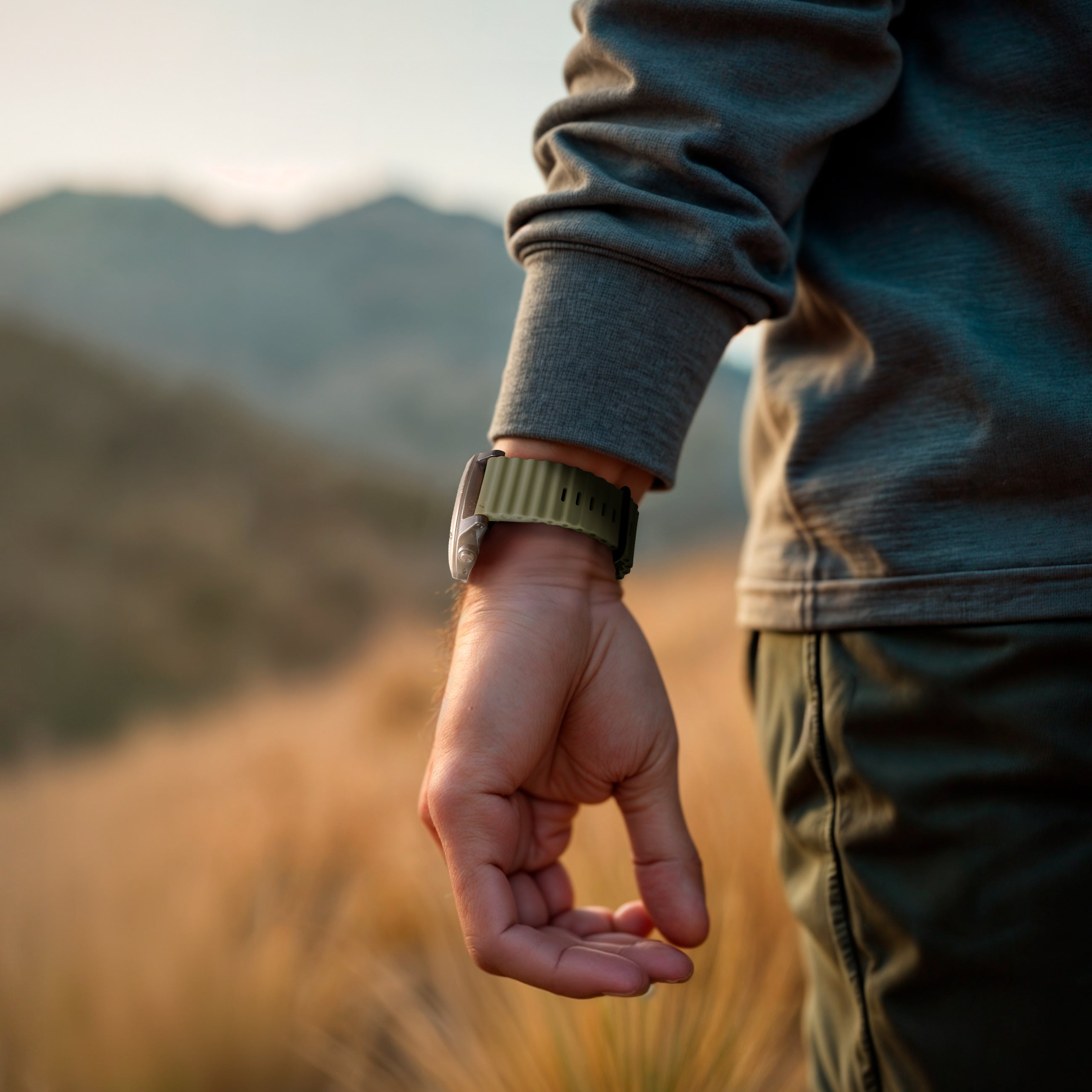 Ein Arm mit einer Uhr in einem Feld. Die Uhr hat ein grünes Armband. Die Person trägt ein graues Hemd und eine grüne Hose.
