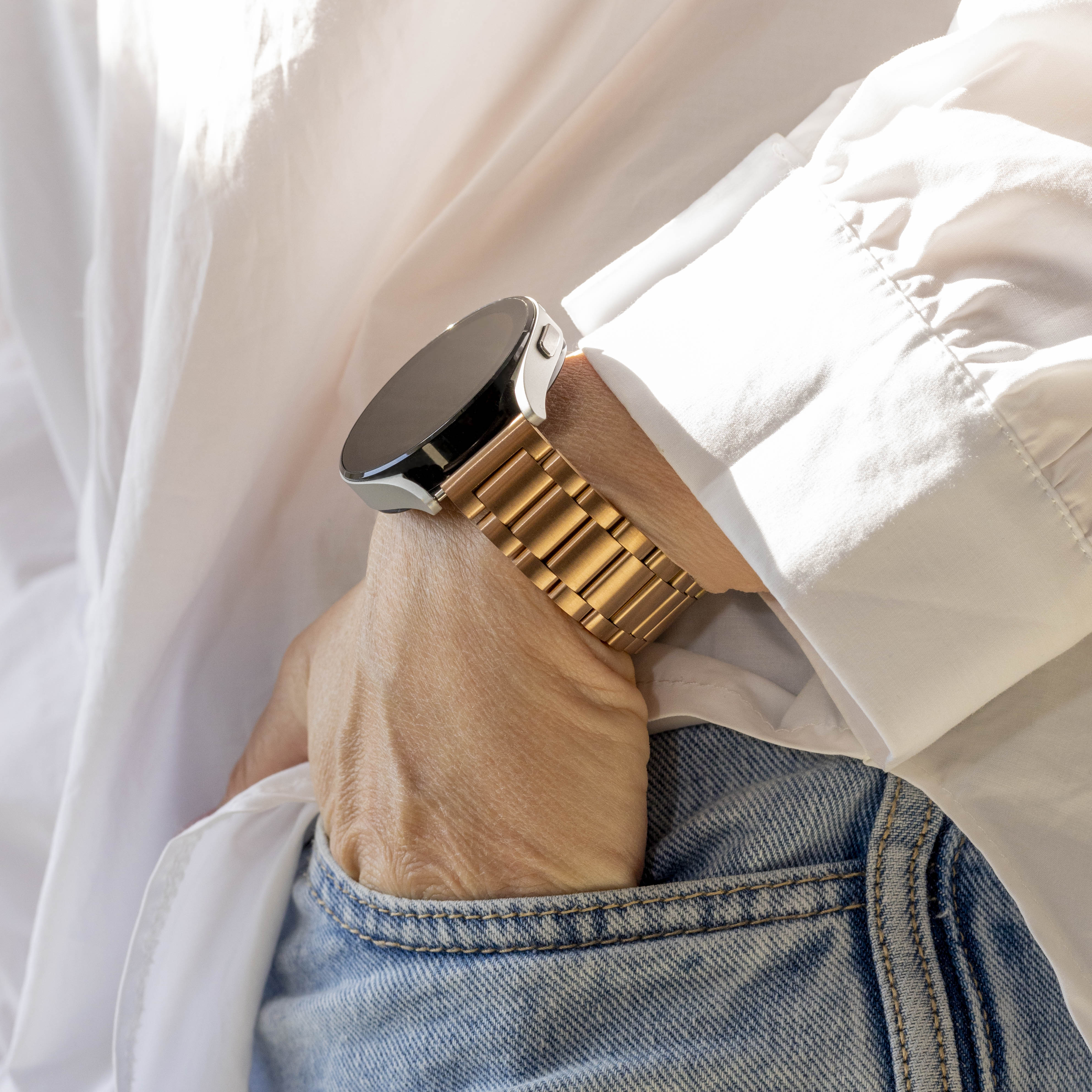 Eine Person mit einer goldenen Uhr am Handgelenk wird gezeigt. Sie trägt ein weißes Hemd und Jeans. Die Uhr ist im Fokus.