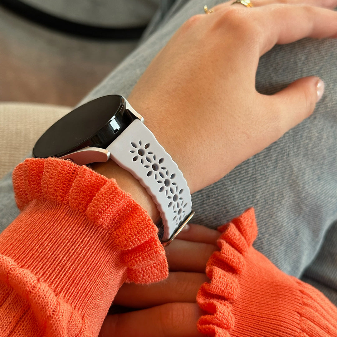 Das Handgelenk einer Person mit einer Smartwatch und einem dekorativen weißen Armband. Die Person trägt einen orangefarbenen Pullover und Jeans.