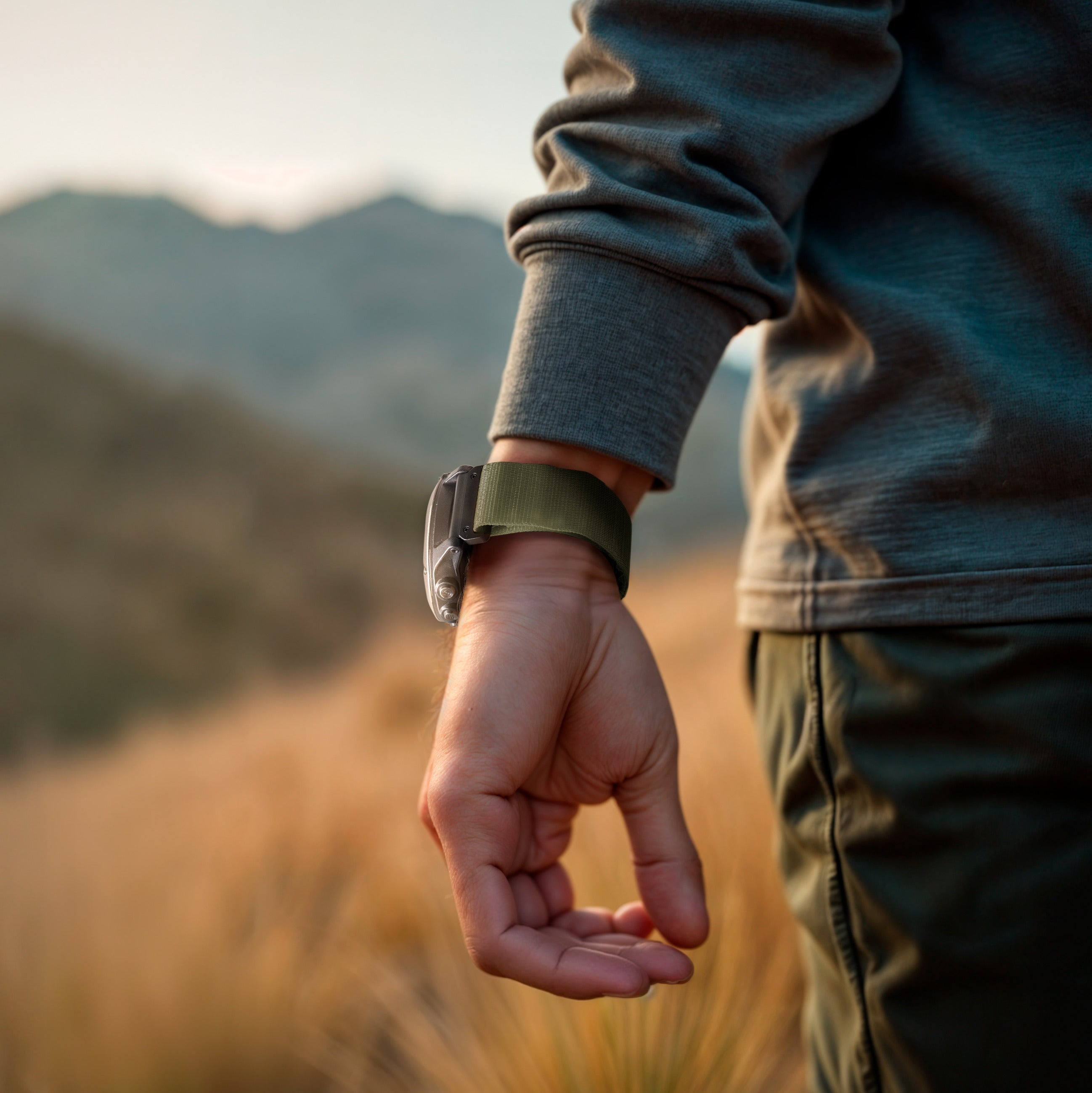 Handgelenk mit Smartwatch und olivgrünem Armband. Im Hintergrund ein Hügel mit trockenem Gras und Bergen.